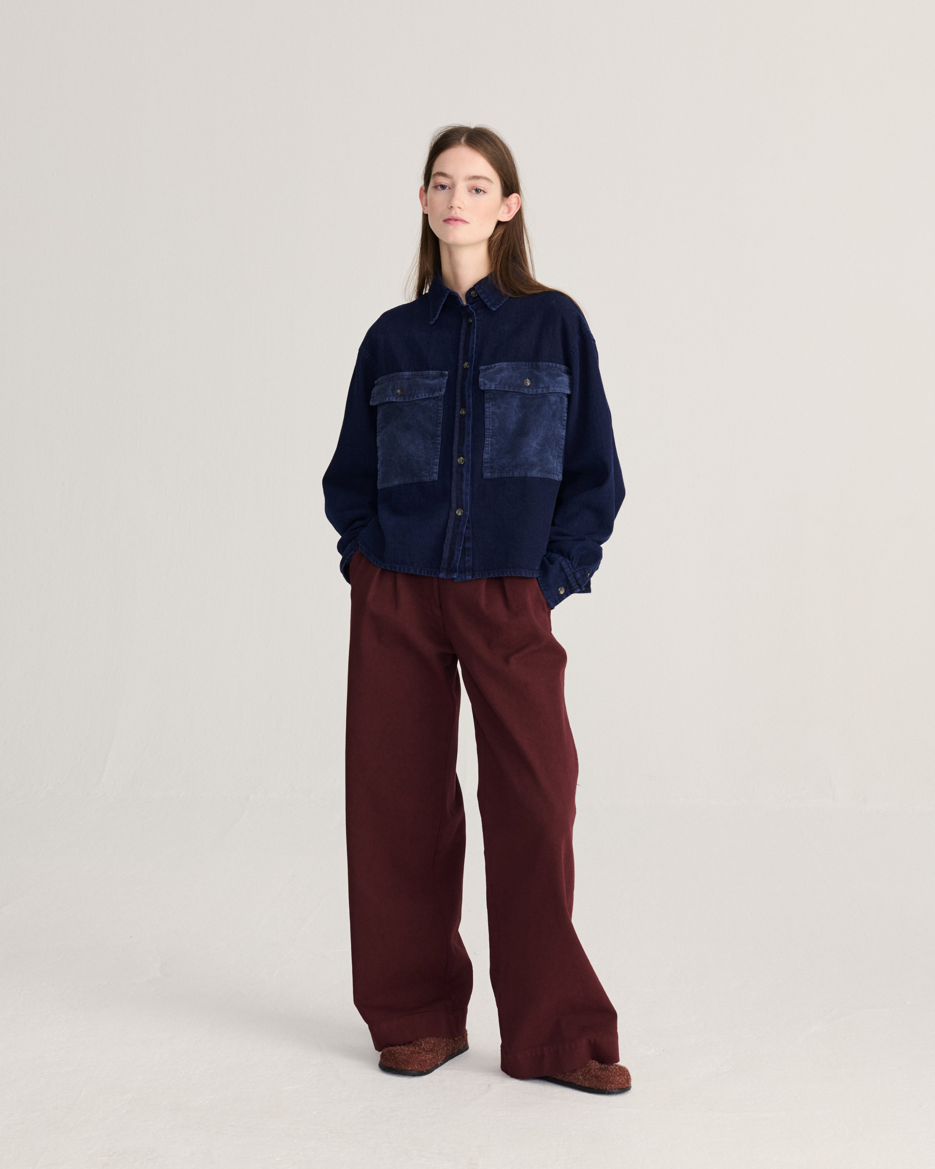 A model stands against a light gray studio background wearing a dark navy patchwork button-up shirt with wide-leg burgundy pants and brown shoes.