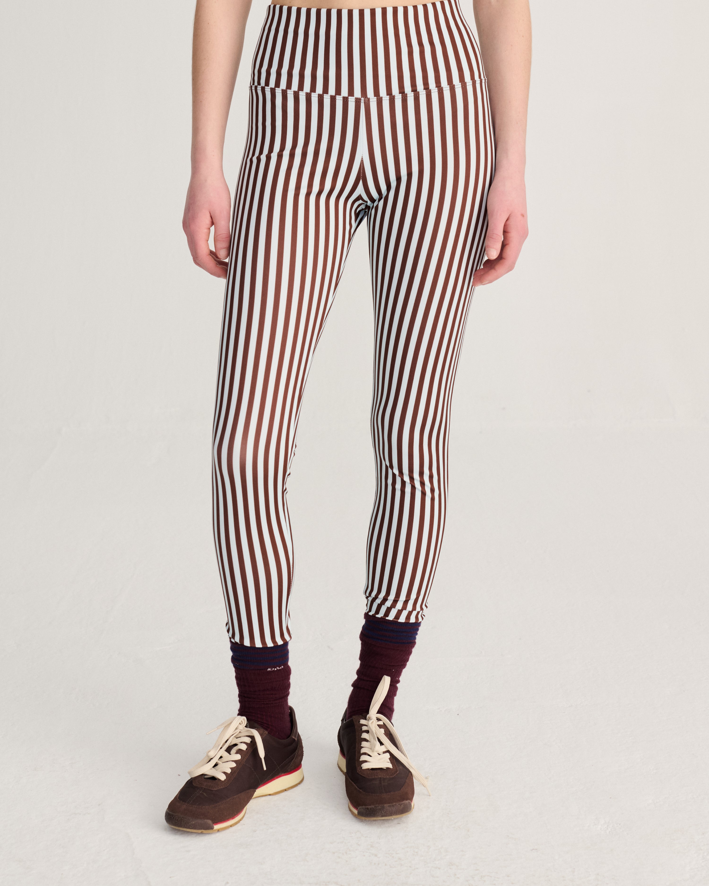 A model from the waist down wearing high-waisted leggings with vertical brown, white, and black stripes, styled with dark maroon socks and brown sneakers against a light gray studio background.