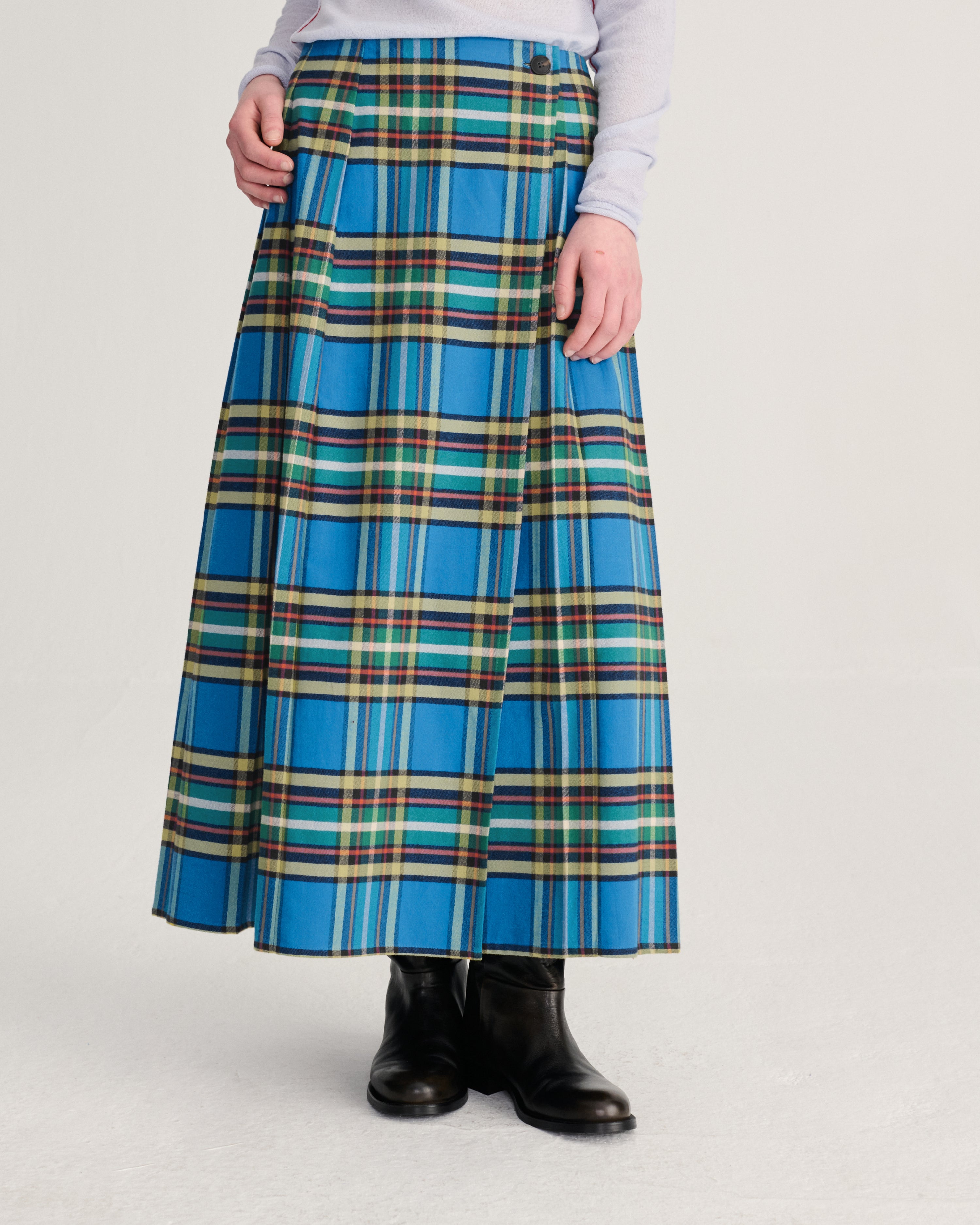A model wearing a high-waisted, pleated A-line skirt in a vibrant blue tartan plaid pattern with green, yellow, and white stripes, styled with a light gray top and black leather ankle boots against a light gray studio background.