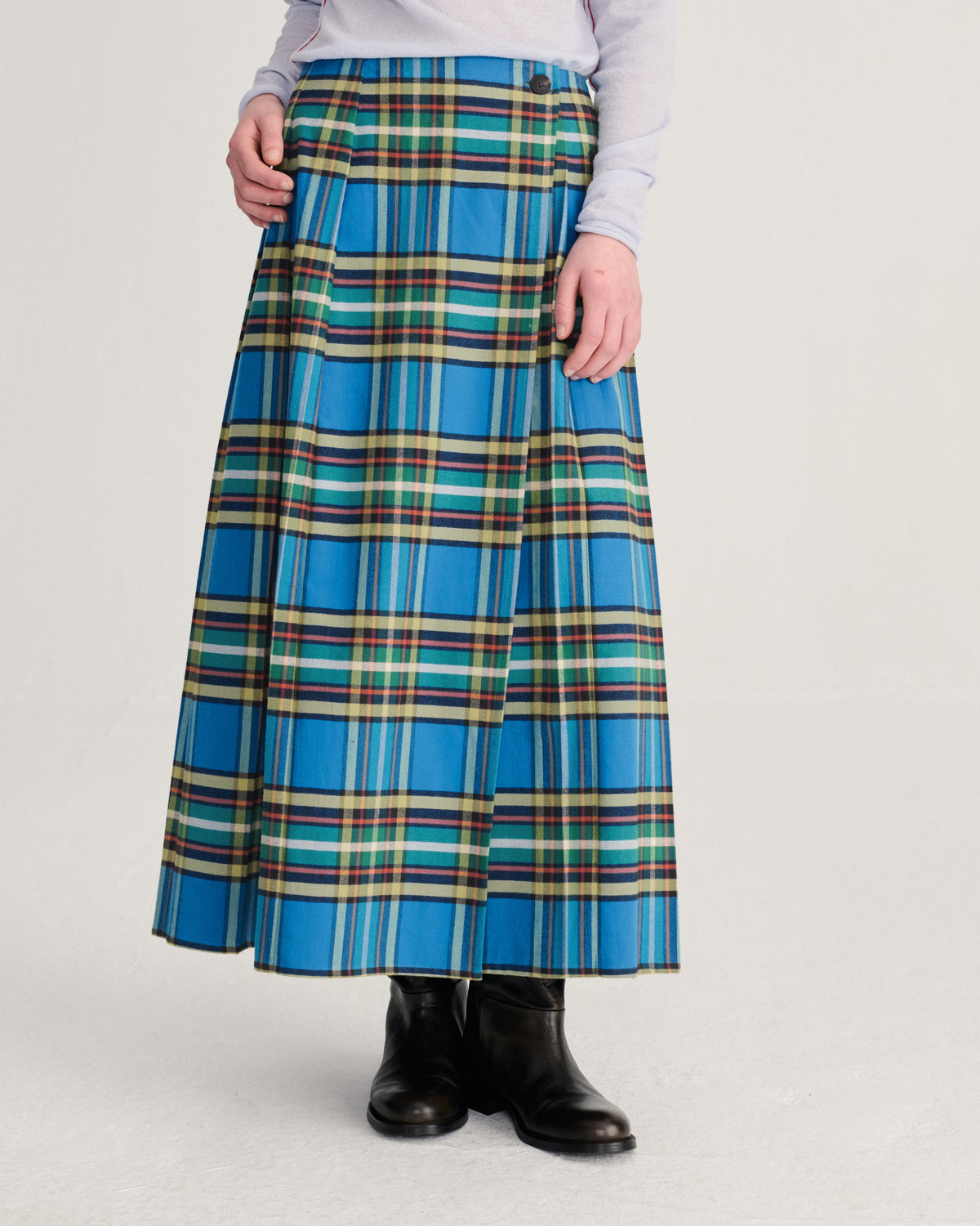 A model wearing a high-waisted, pleated A-line skirt in a vibrant blue tartan plaid pattern with green, yellow, and white stripes, styled with a light gray top and black leather ankle boots against a light gray studio background.