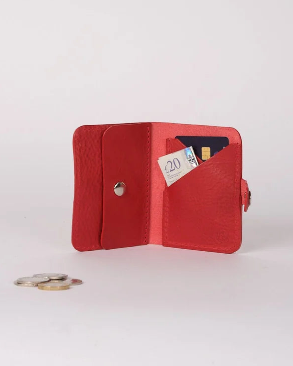A small red leather bifold wallet shown open on a light gray studio background, with card slots holding cards and a coin placed nearby.