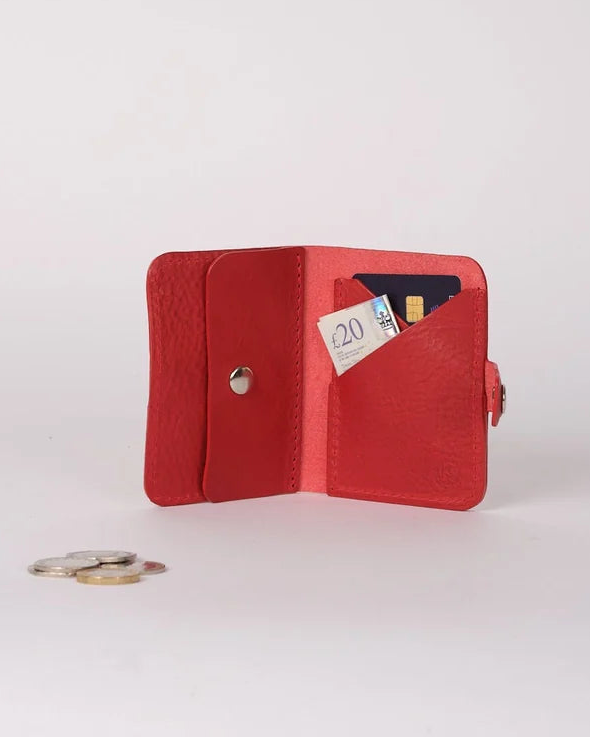 A small red leather bifold wallet shown open on a light gray studio background, with card slots holding cards and a coin placed nearby.
