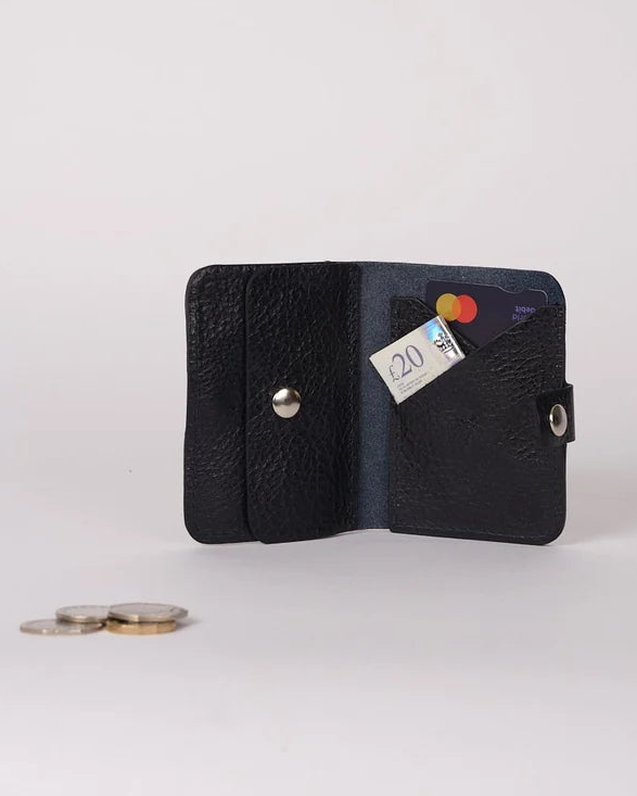 A small black textured leather wallet shown open on a light gray studio background, with a card partially inserted and a few coins placed beside it.