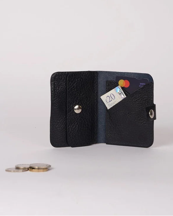 A small black textured leather wallet shown open on a light gray studio background, with a card partially inserted and a few coins placed beside it.