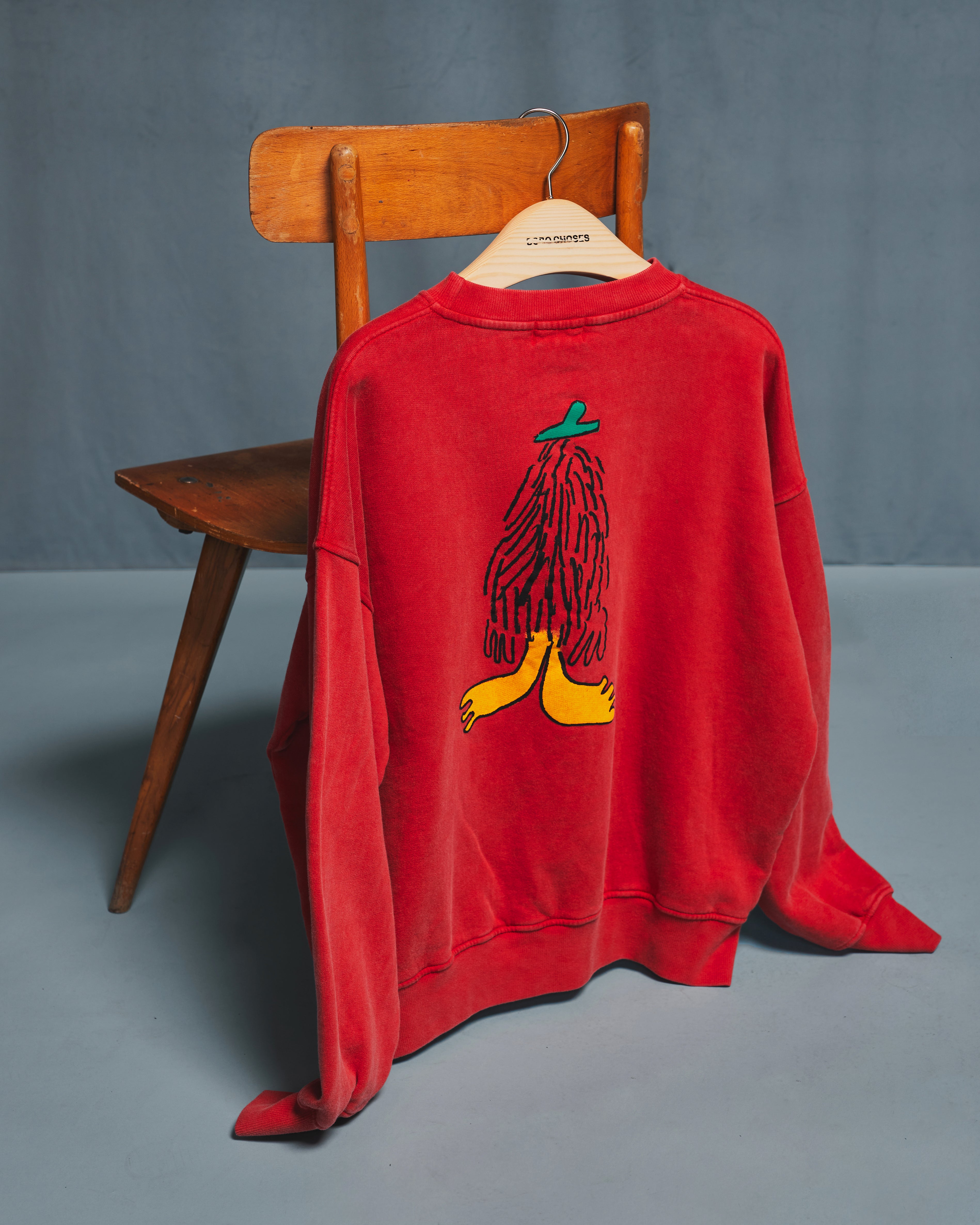 A red crewneck sweatshirt hanging on a wooden hanger from a wooden chair, showing a graphic of a figure with black hair and a green hat on the back, set against a gray studio background.