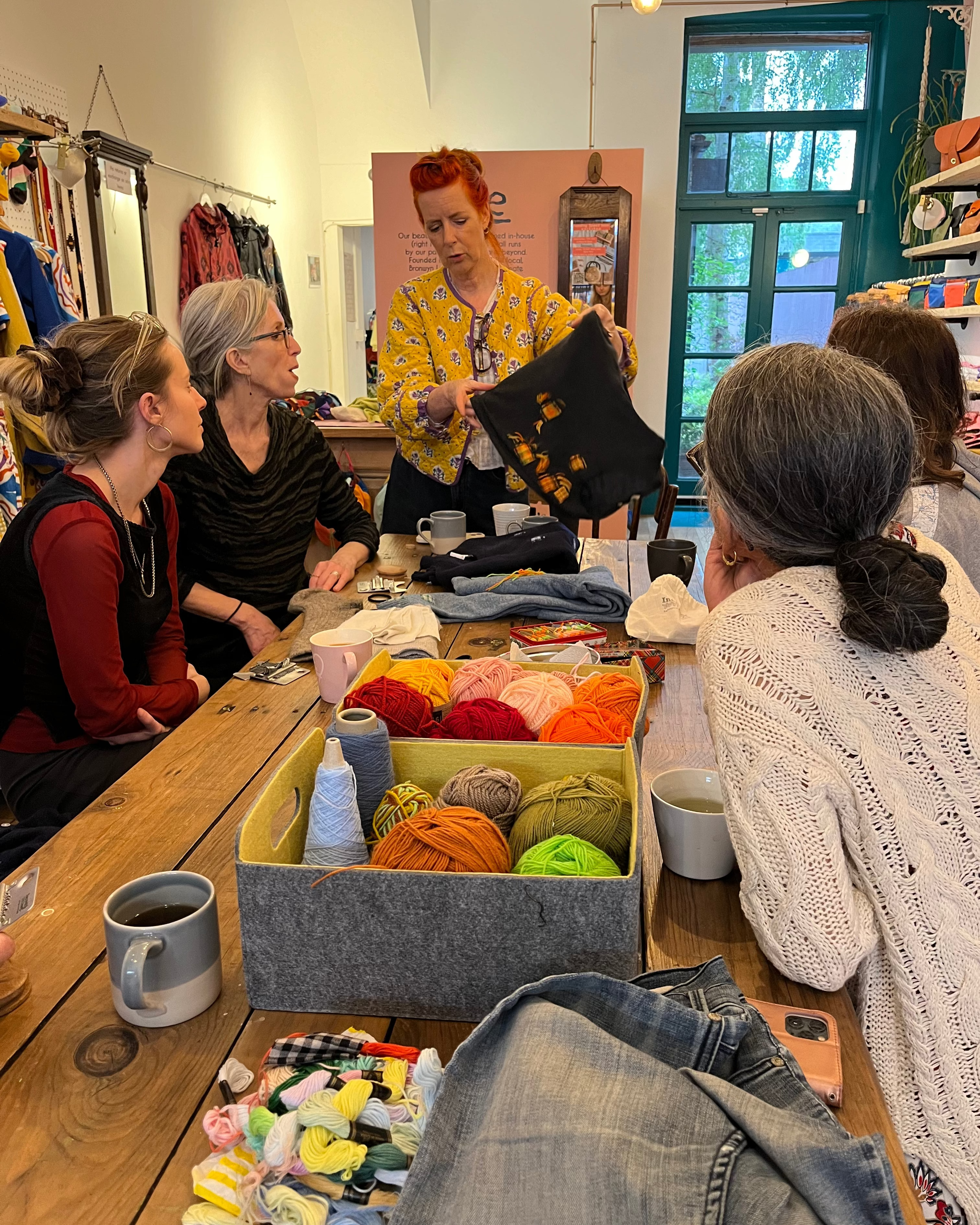 Group of people at a craft workshop with yarn and knitting materials on a table.