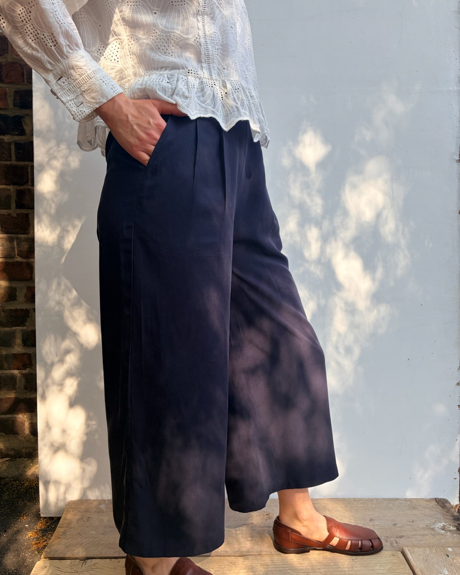 A person wearing navy wide-leg cropped trousers, a white lace-trim blouse, and brown leather loafers, standing outdoors against a white wall with dappled sunlight and shadows.