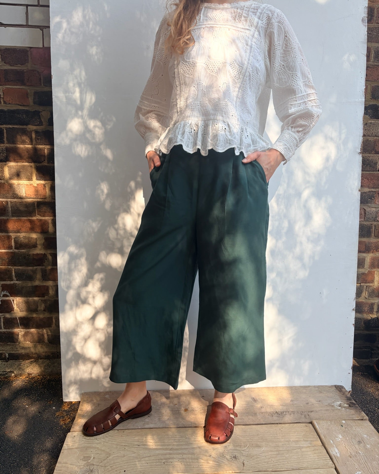 A person wearing a white long-sleeve lace-trim blouse and dark green wide-leg cropped pants, standing outdoors against a white wall with brick sides and dappled sunlight.