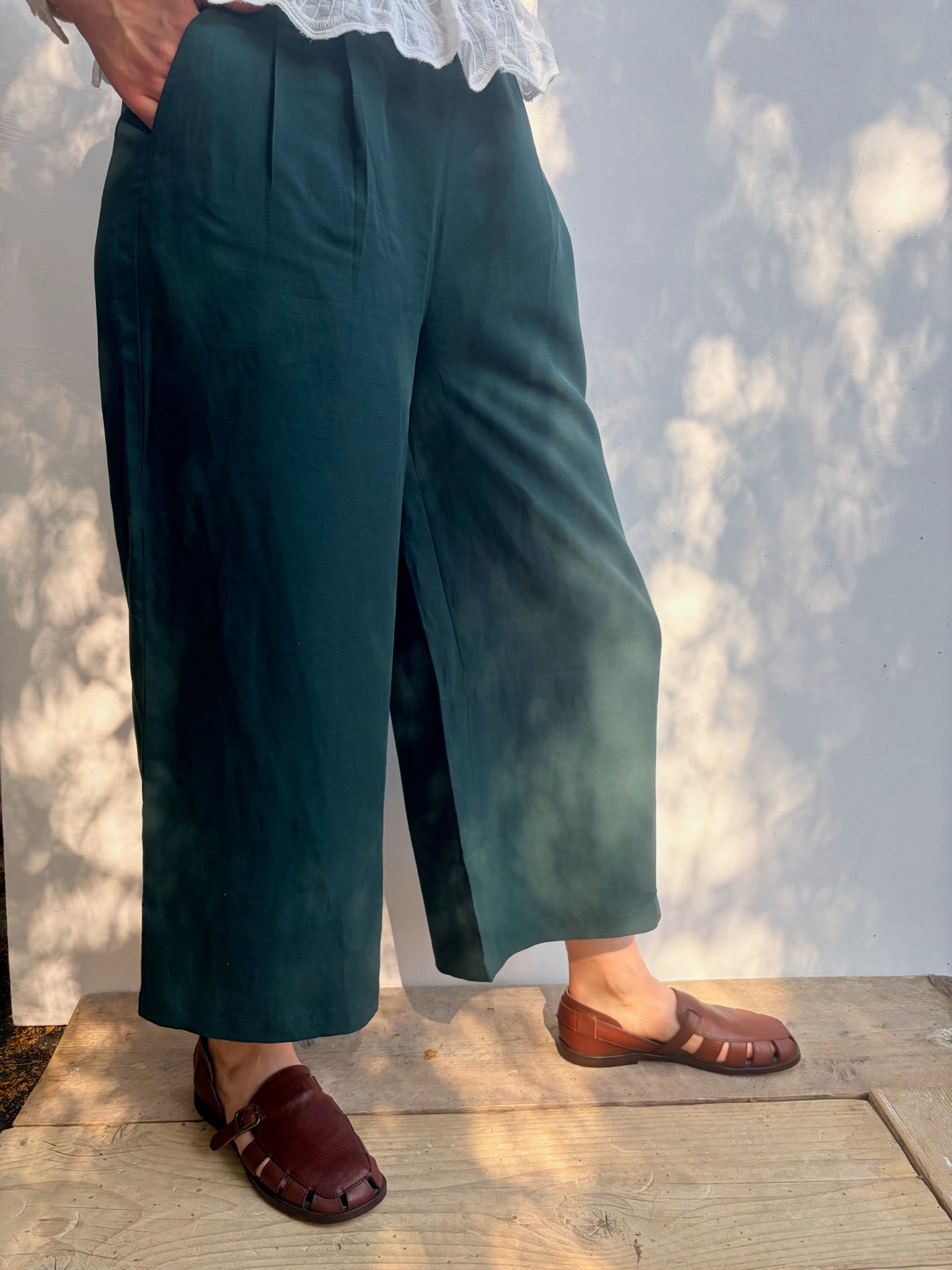 A person wearing dark teal wide-leg cropped pants with pleats, paired with brown leather slide sandals and a white lace-trim top, standing on a wooden deck against a wall with dappled sunlight.