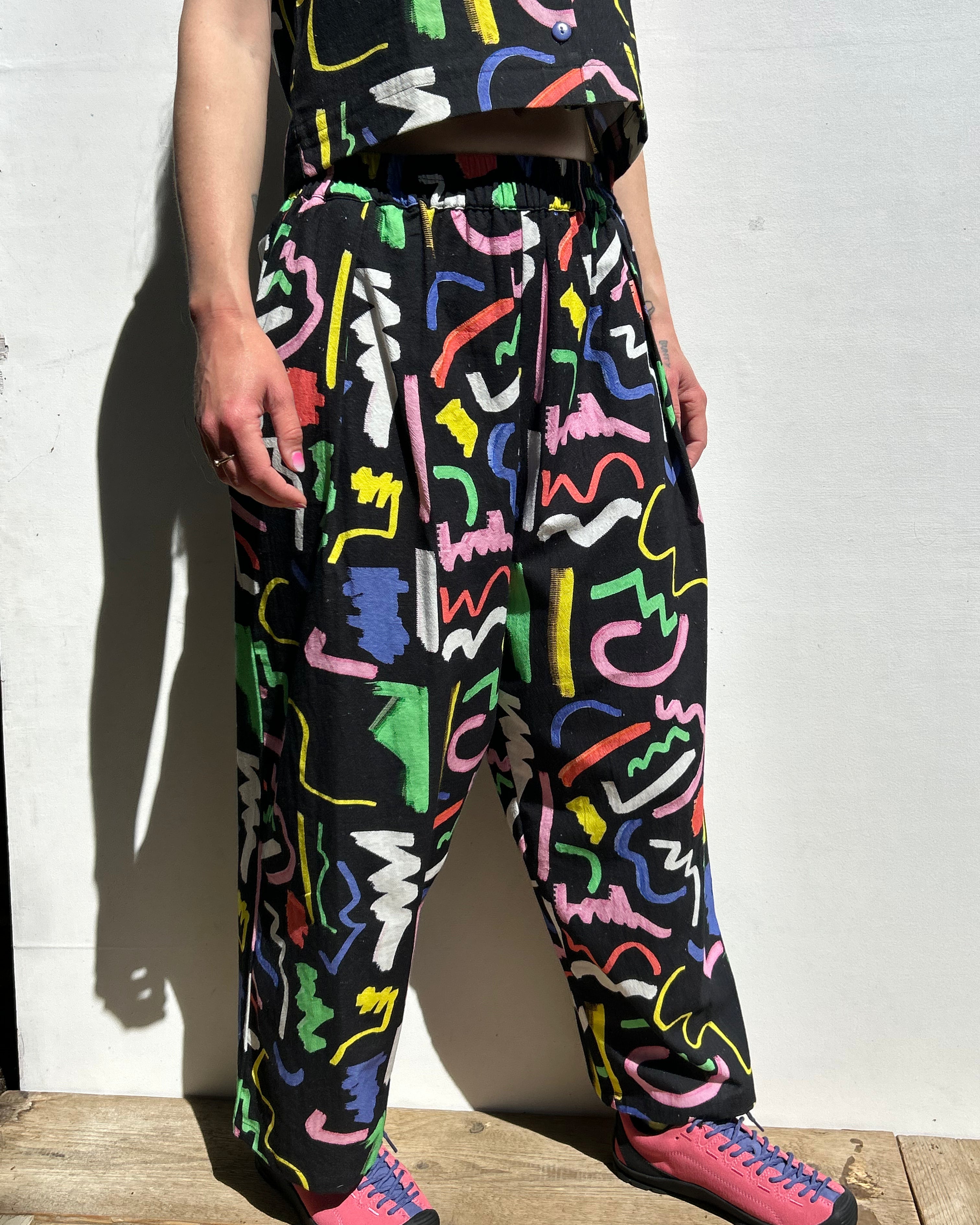 Person wearing colorful abstract pattern pants and pink shoes against a white wall.