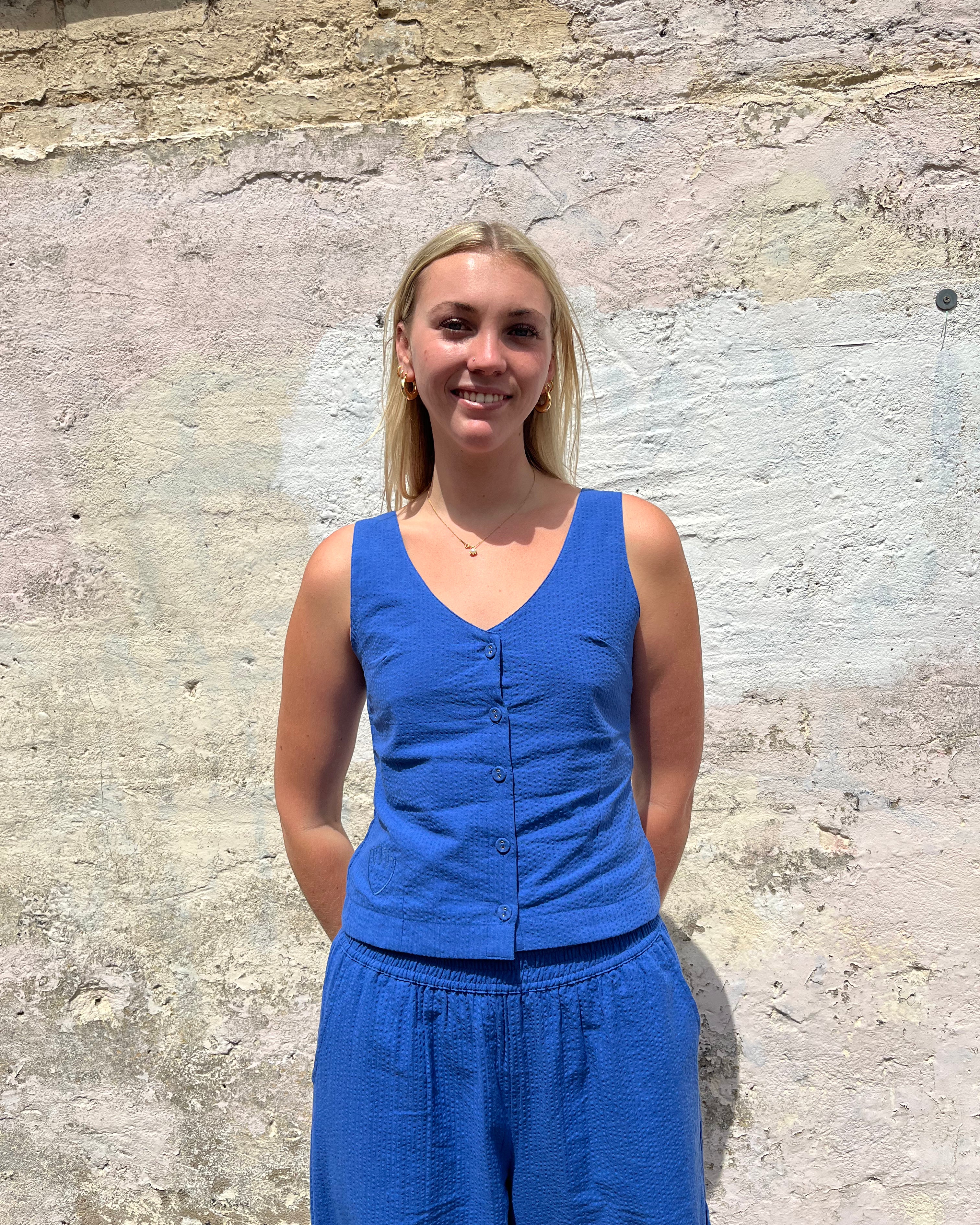 Woman wearing a blue sleeveless top and pants standing against a stone wall.