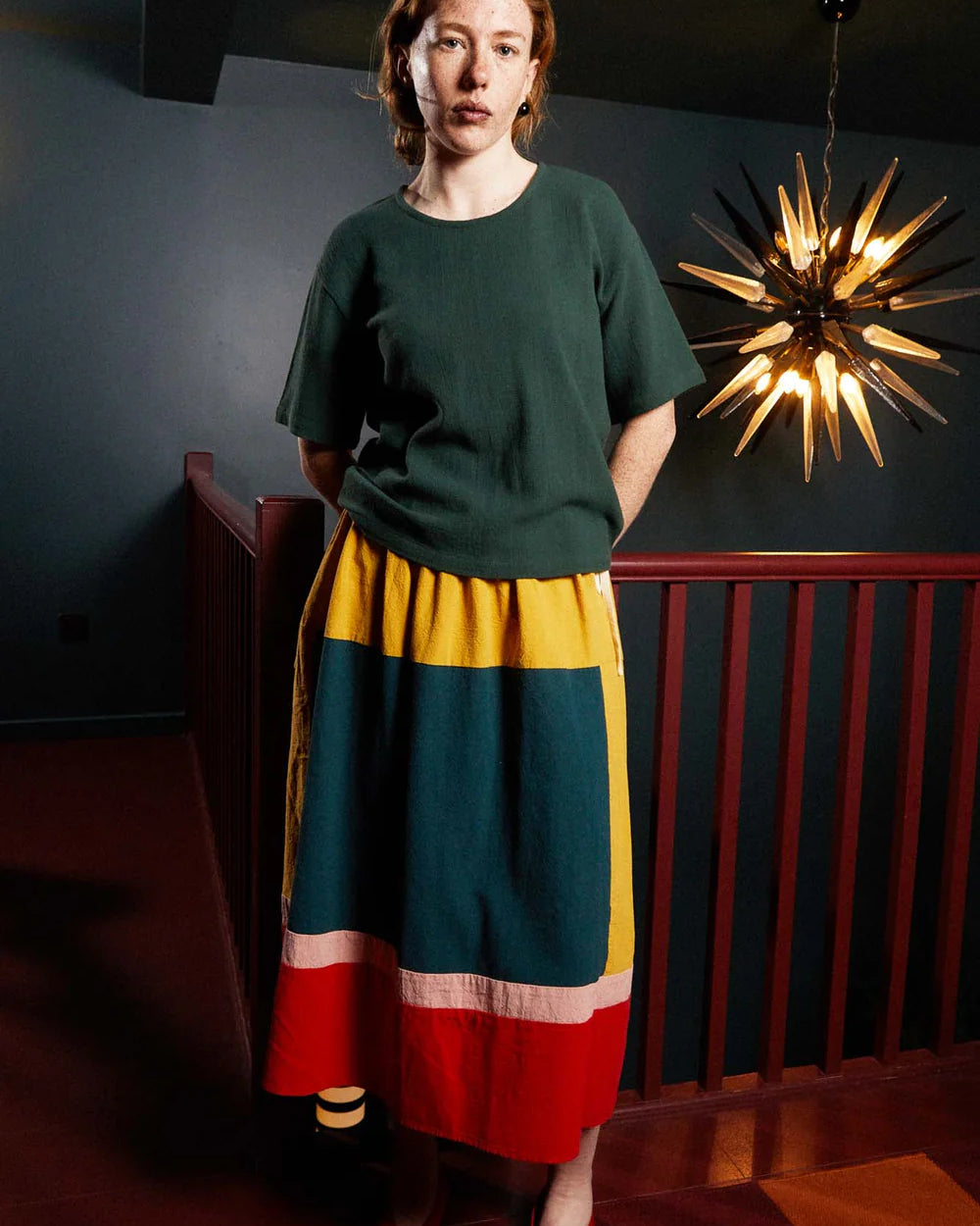 Woman wearing a green top and colorful skirt standing in a room with a decorative light fixture.