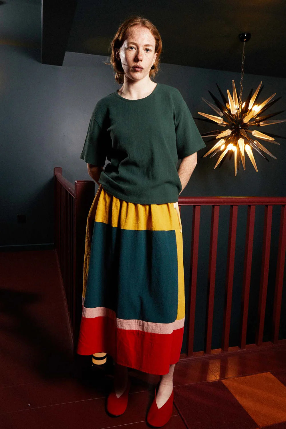 Woman wearing a green top and colorful skirt standing in a room with a decorative light fixture.