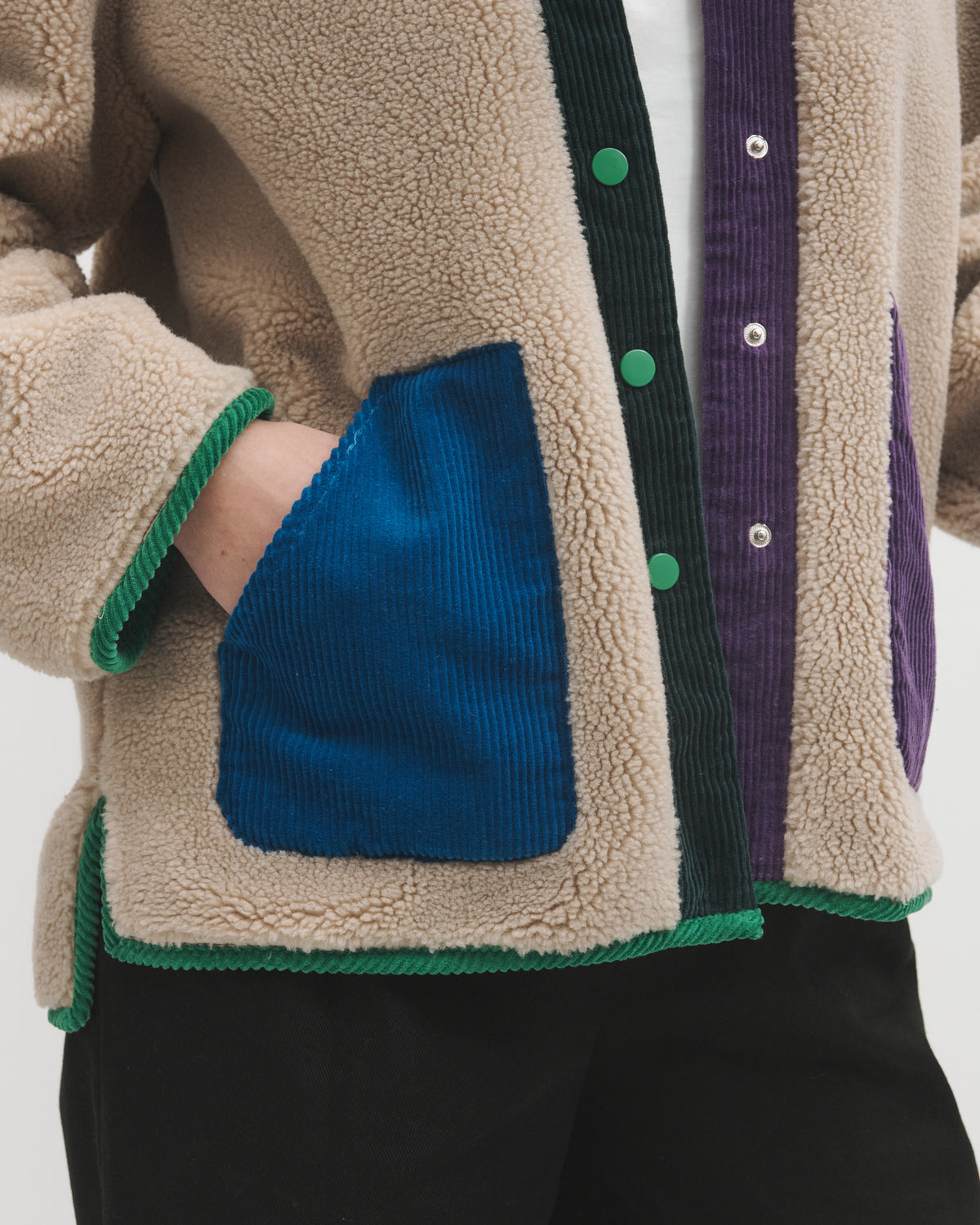 Close-up of a person wearing a beige sherpa cardigan with contrasting green trim, a blue corduroy front pocket, and purple corduroy side panels with green buttons.