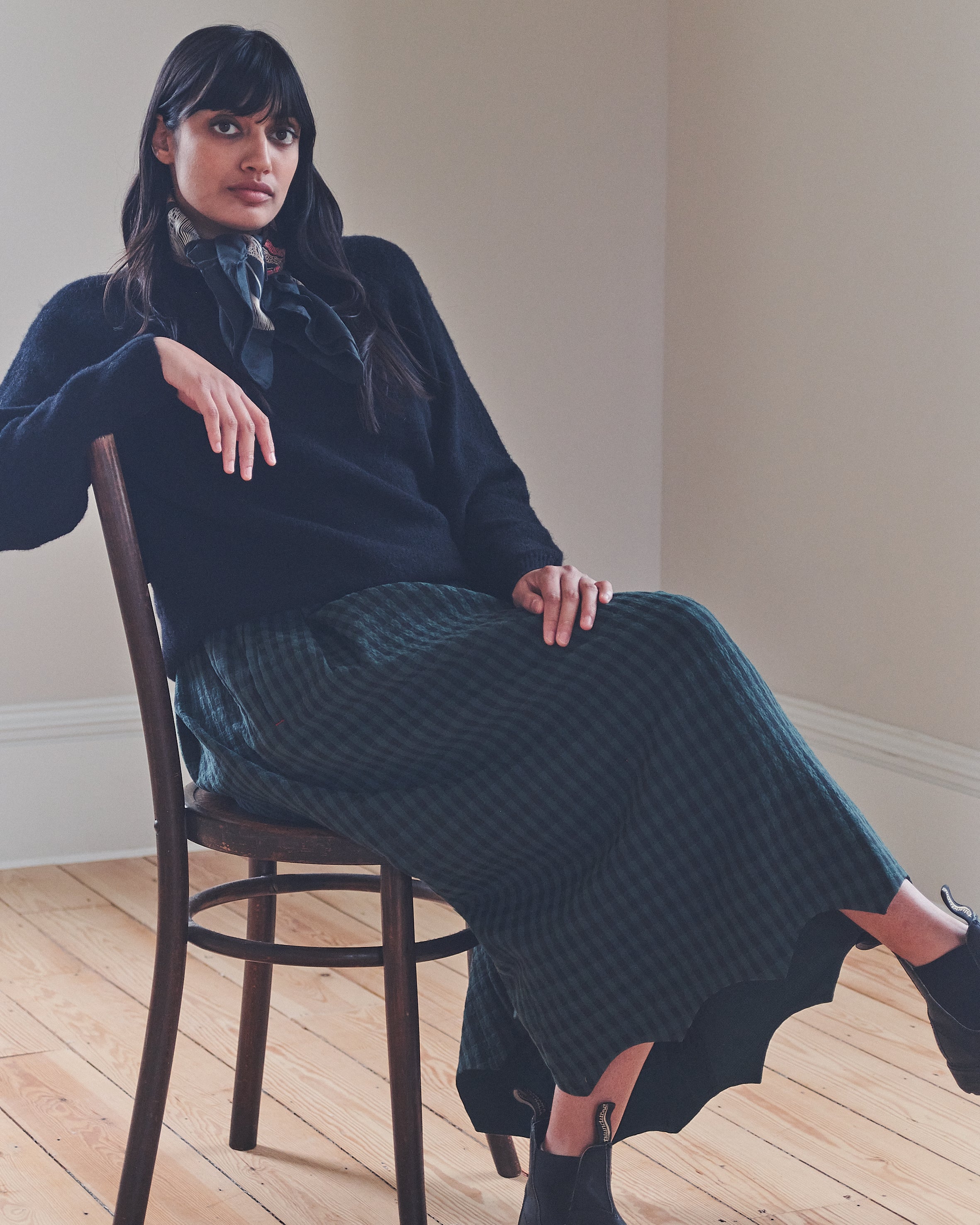 Woman sitting on a chair wearing a dark blue sweater and patterned skirt in a minimalistic room.
