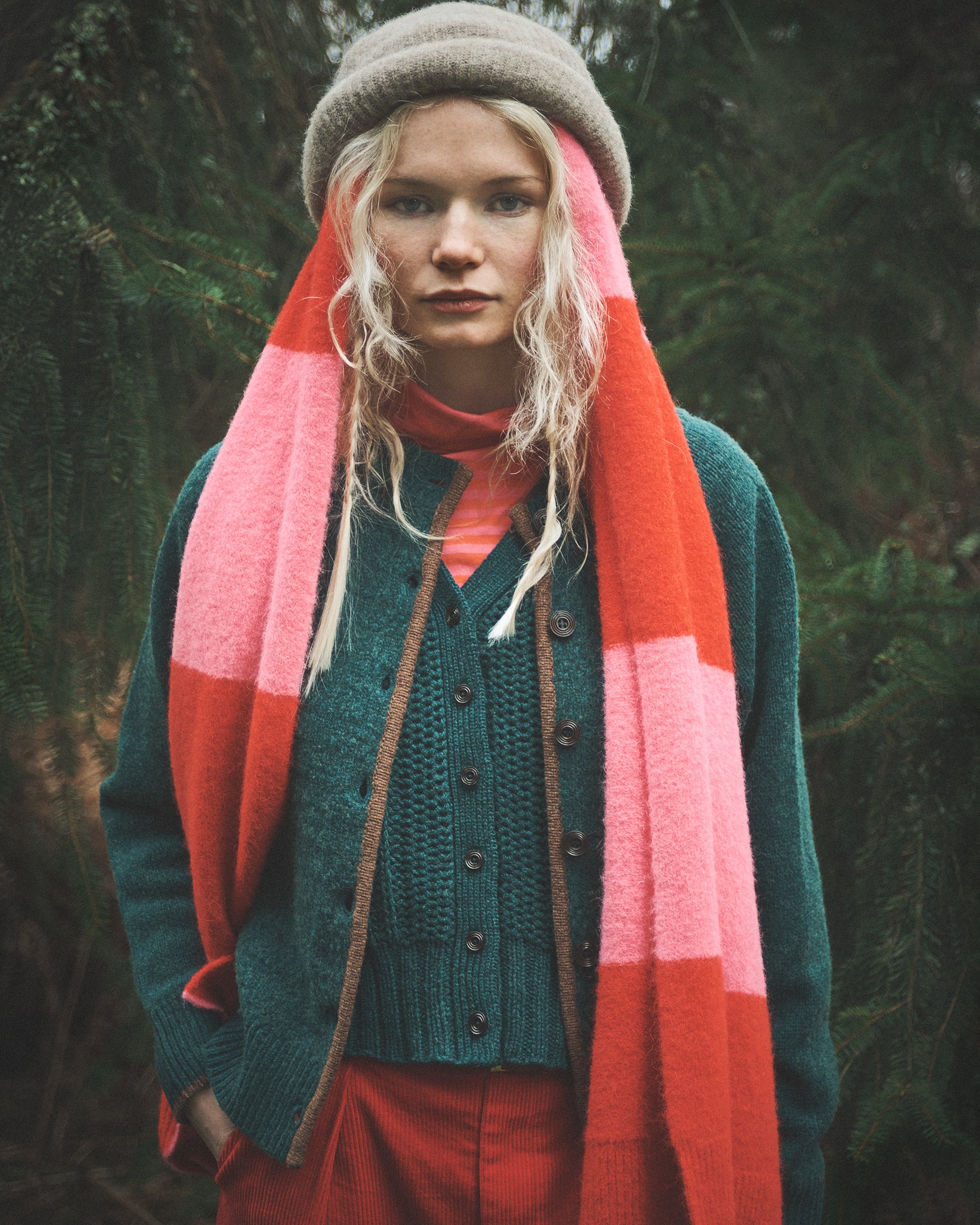 A woman wearing a chunky knit beanie, a teal button-front cardigan, a bright pink and orange striped scarf, and rust-red pants, standing outdoors against a dark forest background.