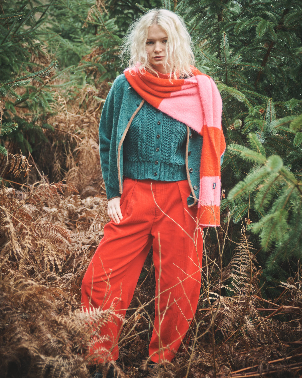 A model stands outdoors in a forest setting wearing a teal knit cardigan, bright red wide-leg pants, a pink and orange striped knit scarf, and a green top.