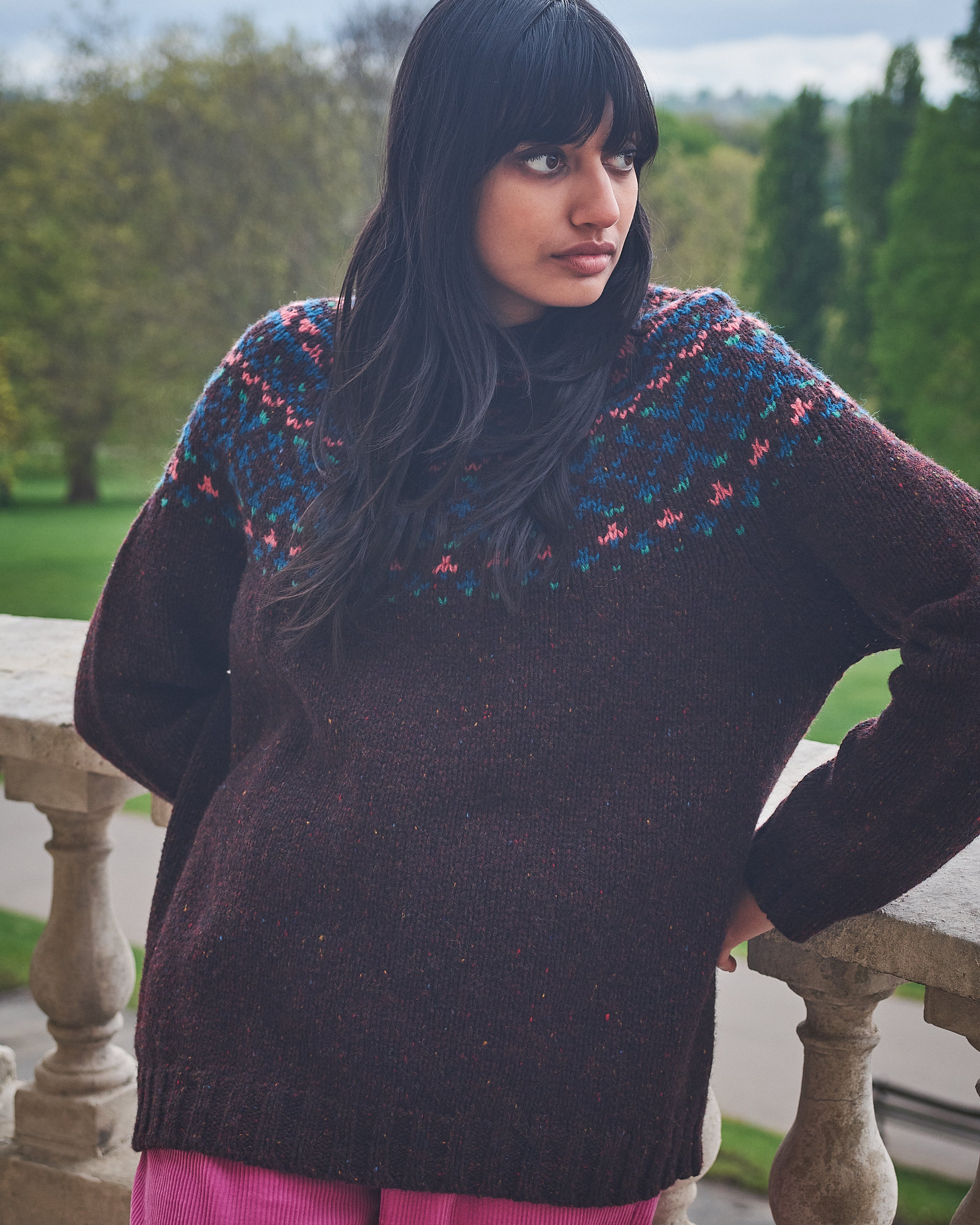 Woman wearing a dark sweater with colorful patterns, standing on a stone balcony with greenery in the background.