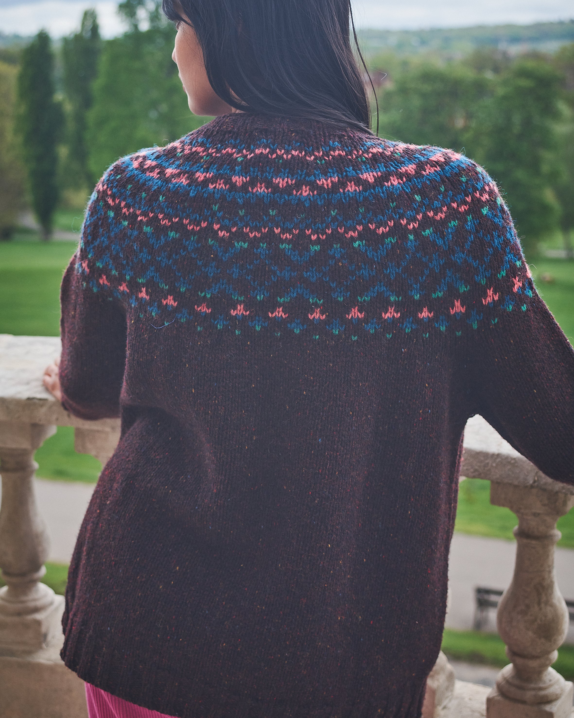 Person wearing a patterned sweater standing on a stone balcony with a scenic view.