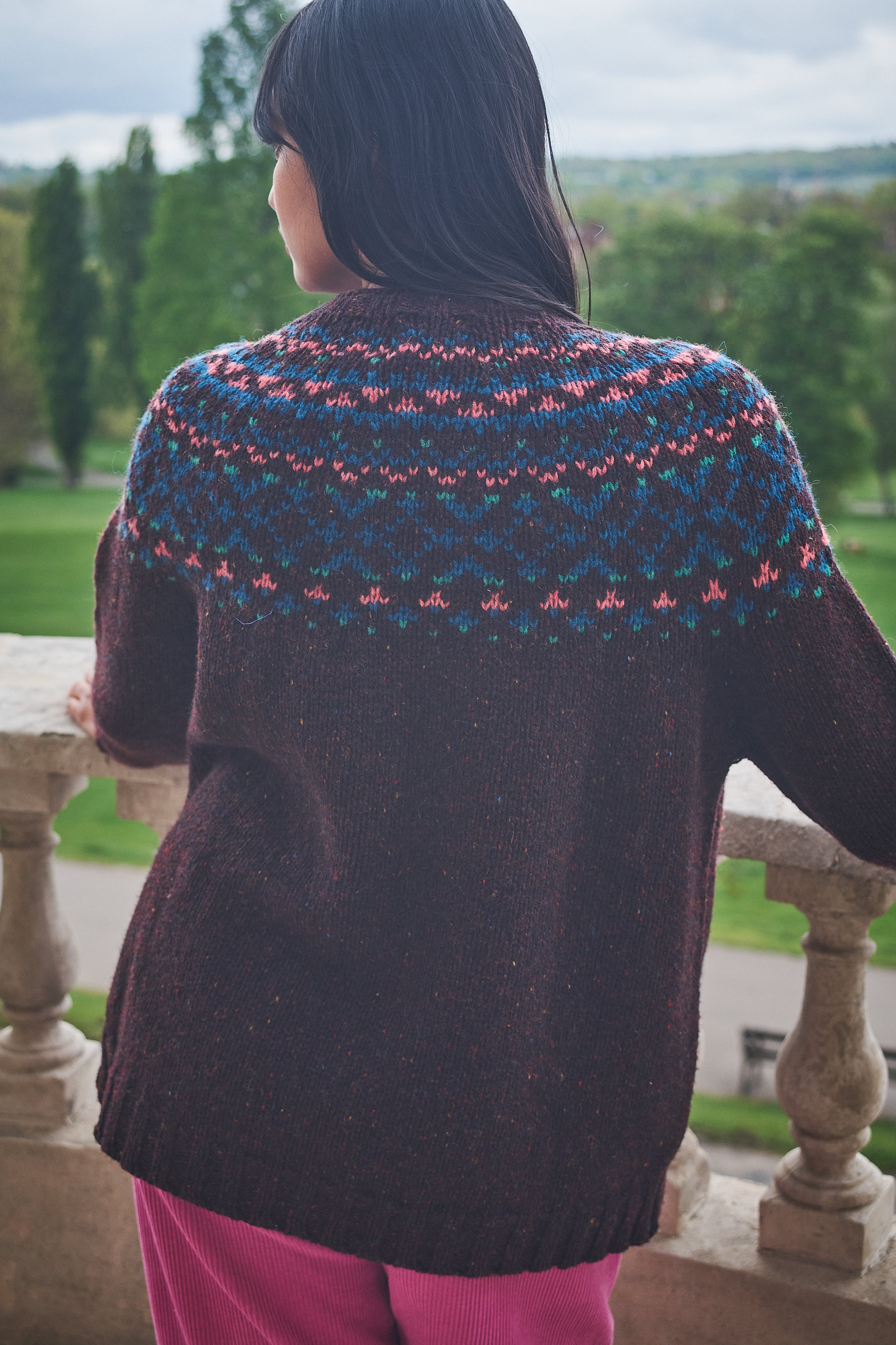 Person wearing a patterned sweater standing on a stone balcony with a scenic view.