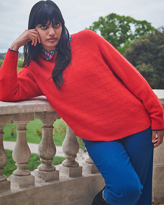 Person wearing a bright red sweater and blue pants standing on a stone railing with greenery in the background