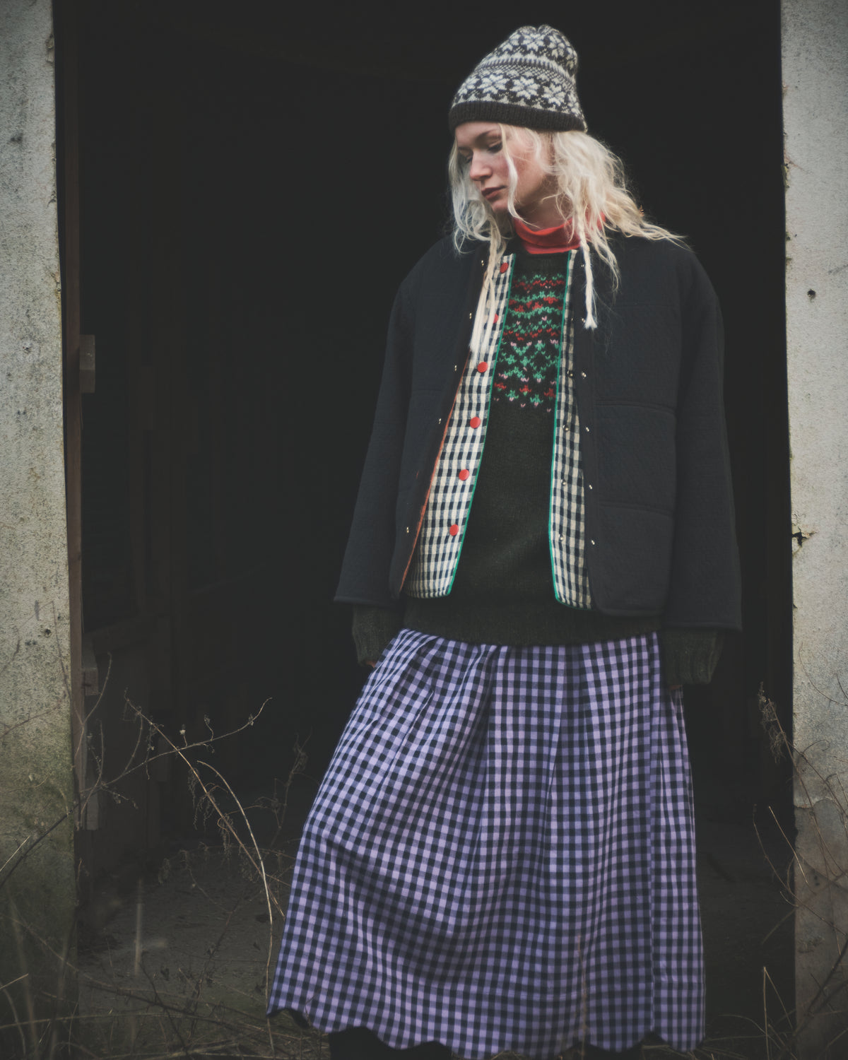 A person standing in a dark doorway wearing a black jacket, a patterned knit beanie, a red and green patterned sweater, and a blue-and-white gingham skirt.