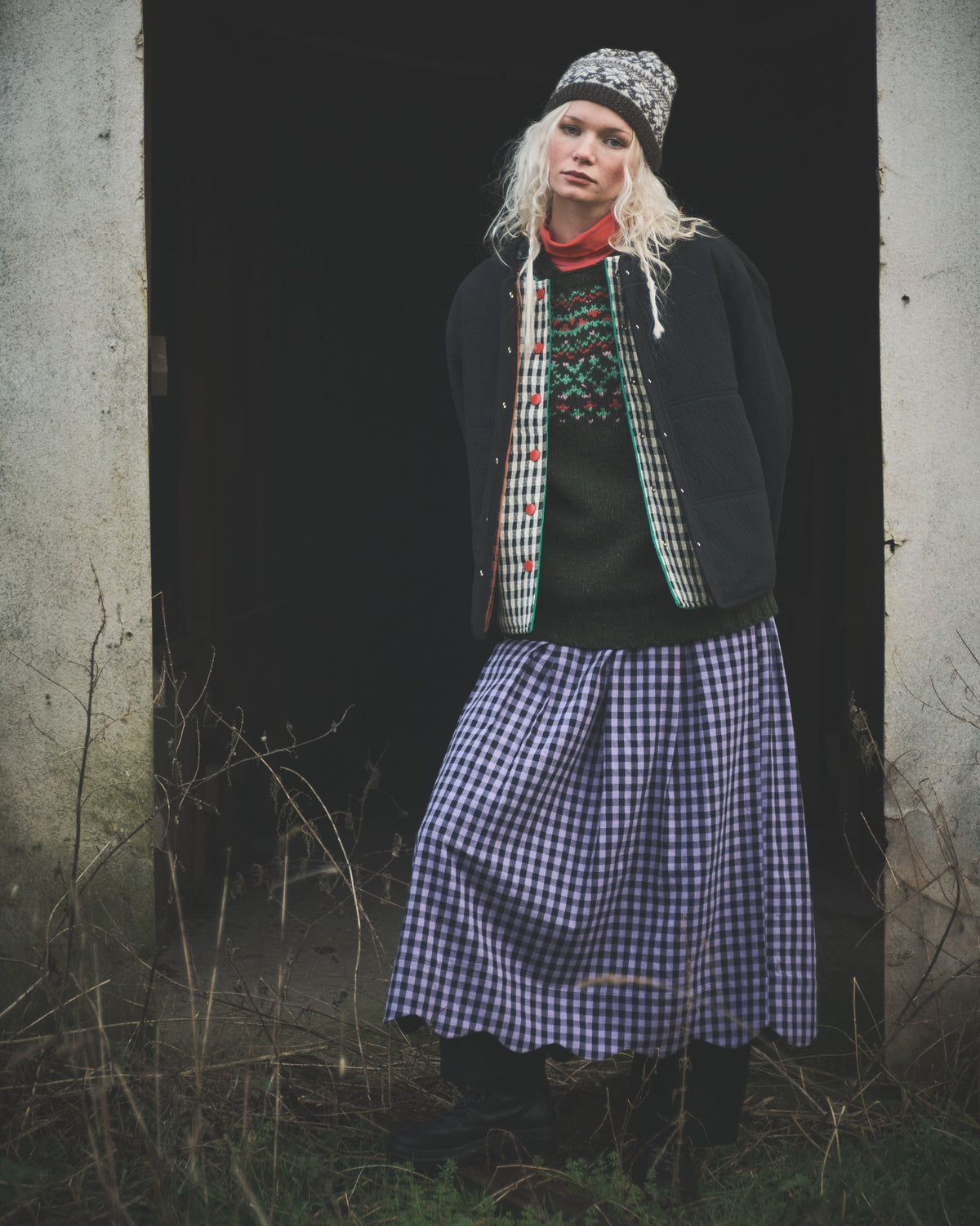 A woman stands in a dark doorway wearing a patterned knit beanie, a dark jacket over a green patterned sweater, a red neck gaiter, and a long blue-and-white gingham skirt.