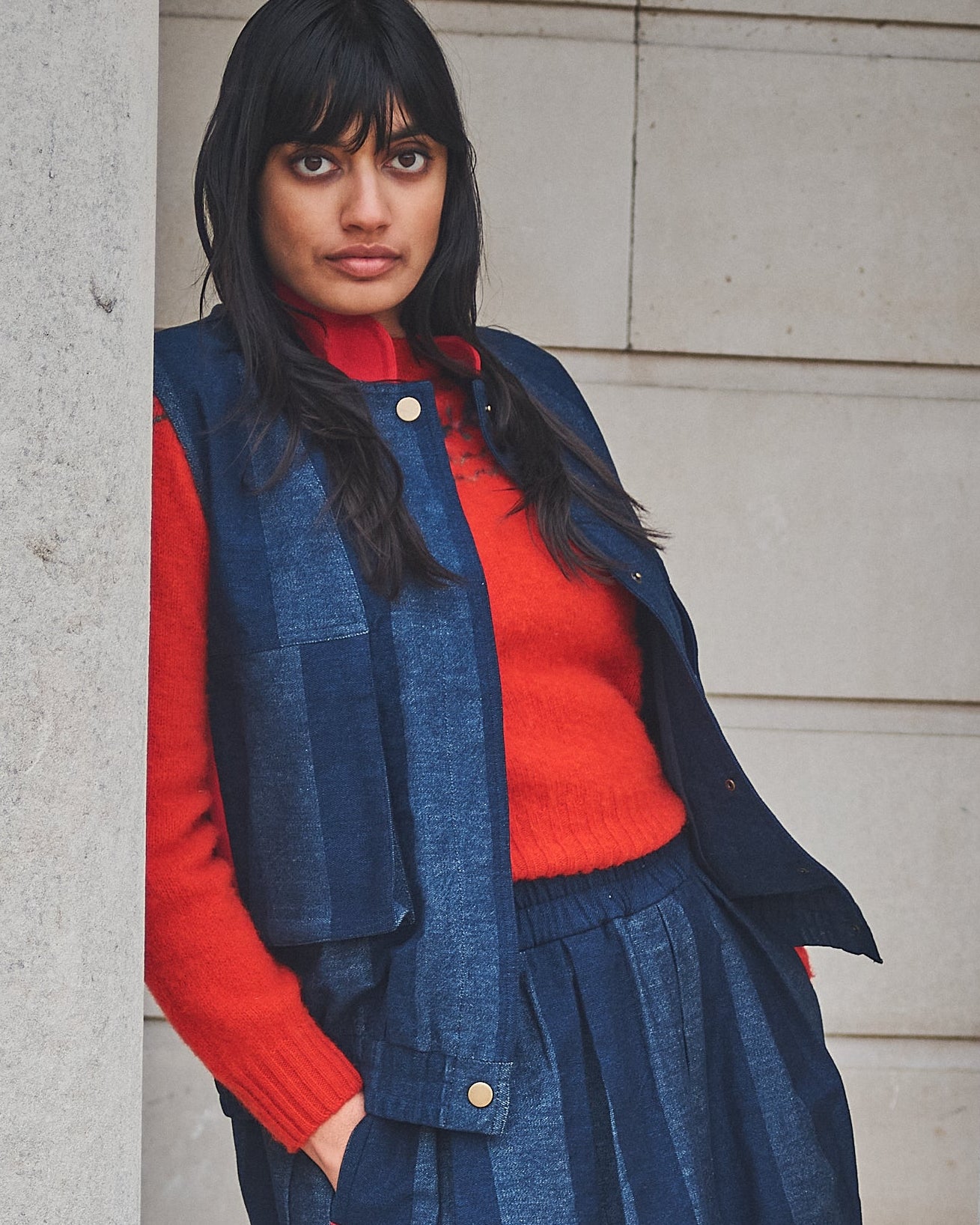 Woman in a Lowie denim stripe gilet, red fair isle jumper and blue skirt standing between stone columns.
