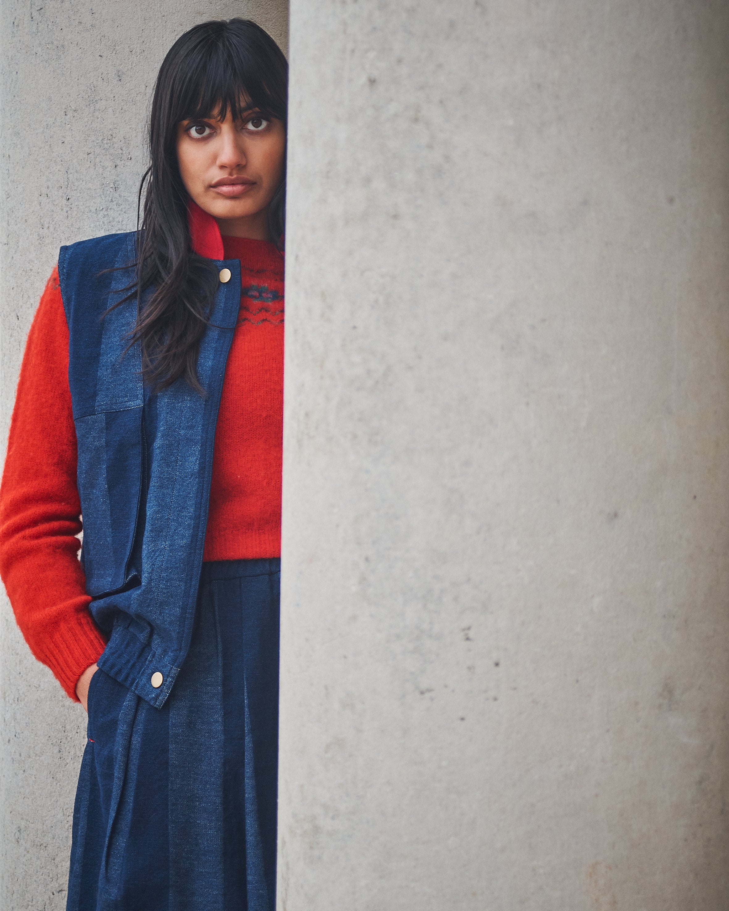Person wearing a red sweater and blue vest against a gray wall