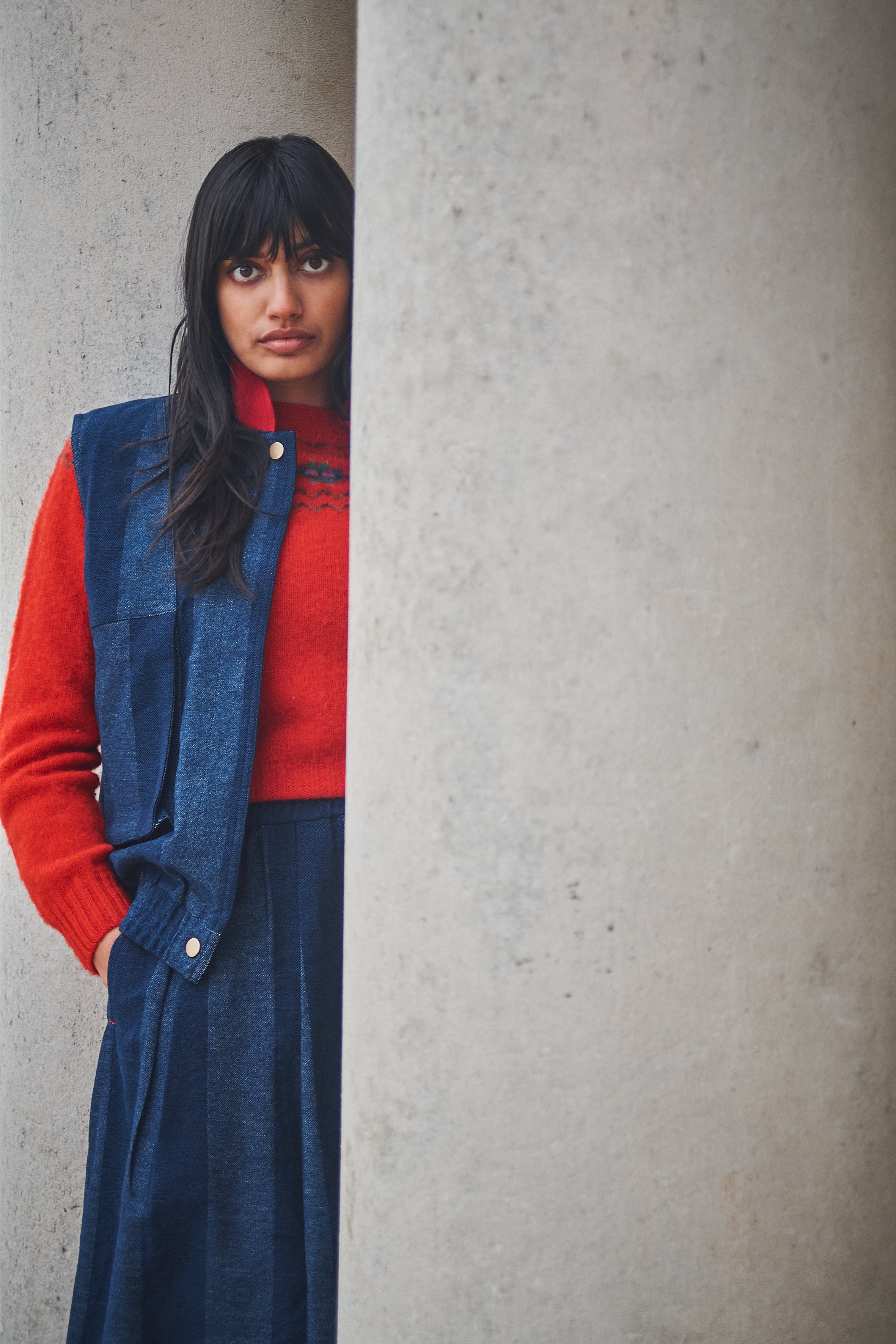 Person wearing a red sweater and blue vest against a gray wall