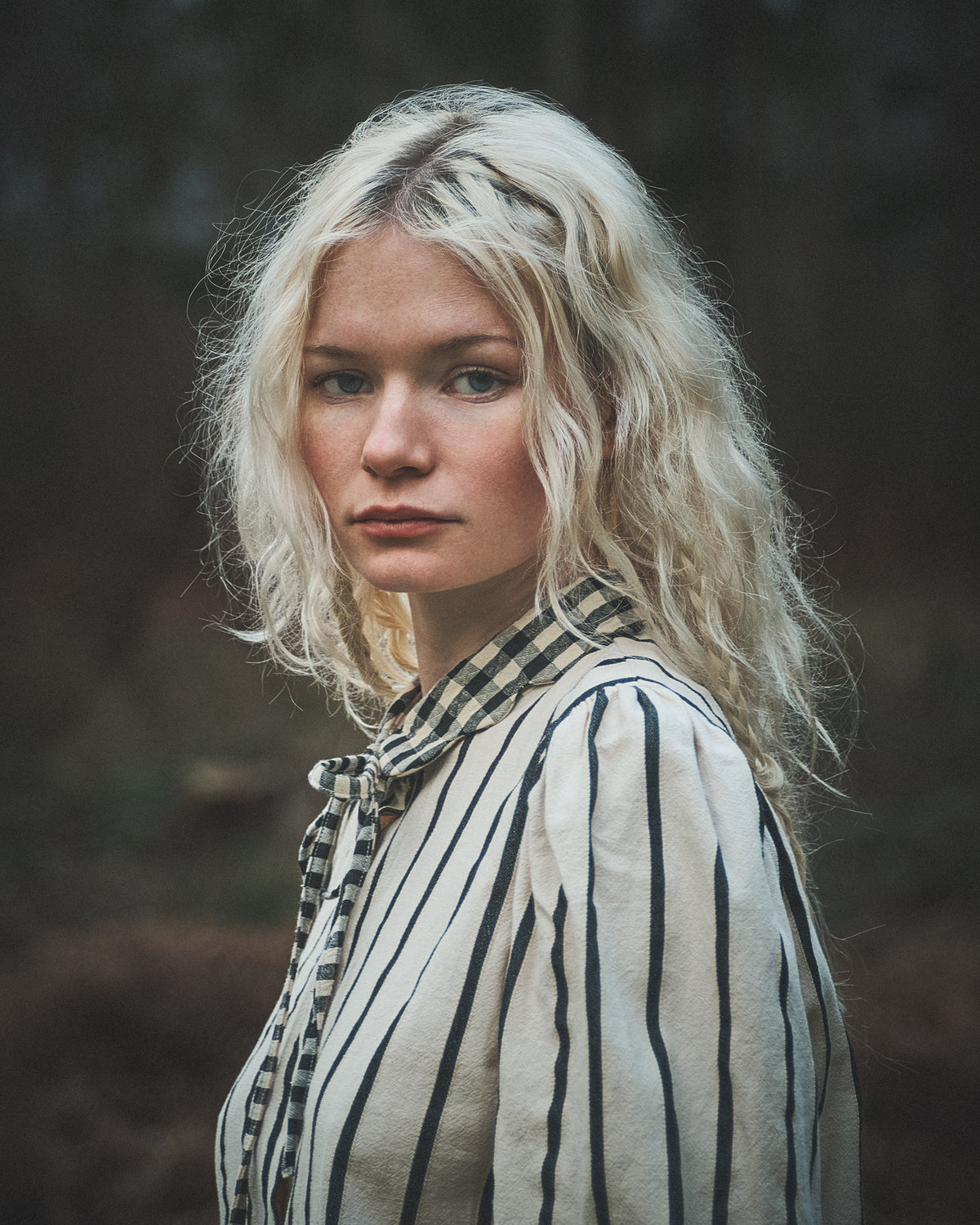 A woman with wavy platinum blonde hair wearing a cream-colored blouse with thin black vertical stripes and a matching striped necktie, photographed outdoors against a dark, blurred forest background.