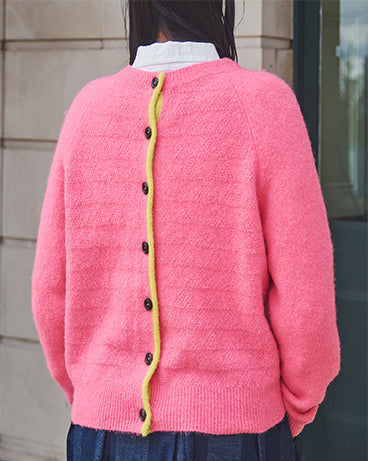 Person wearing a pink cardigan with a yellow trim and black buttons, standing against a neutral background.