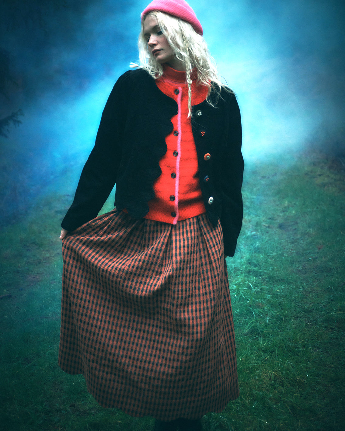 A woman stands outdoors in a misty blue-green environment wearing a bright pink beret, a black cardigan over a vivid orange-red top, and a brown-and-black checkered skirt.