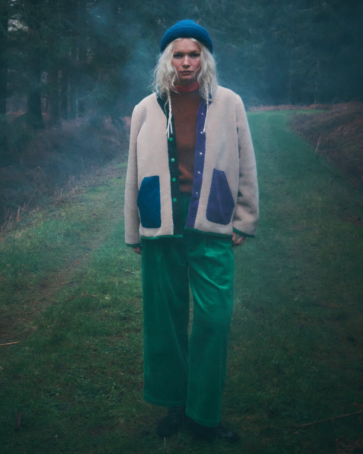 A person standing on a grassy path in a misty forest, wearing a cream cardigan with blue patch pockets, bright green pants, and a blue beret.