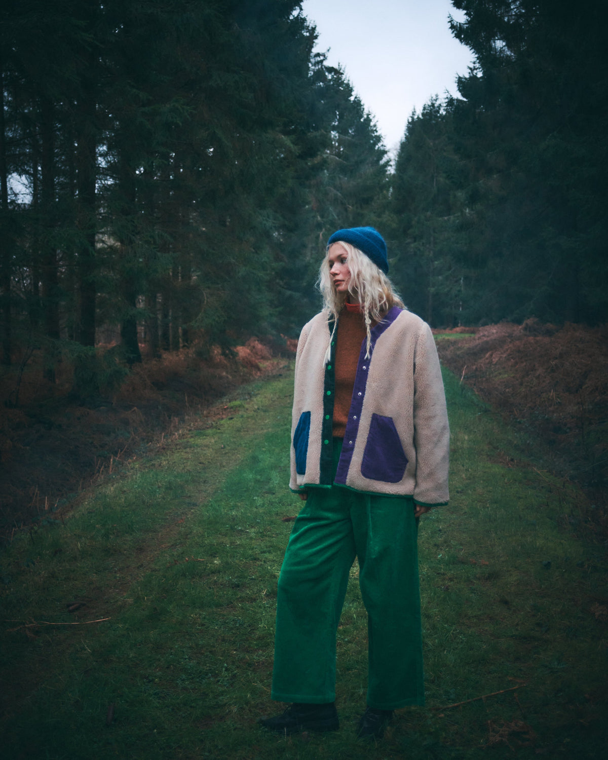 A person standing on a grassy path between dark pine trees, wearing a beige and purple fleece jacket, green wide-leg pants, a brown top, and a blue beanie.