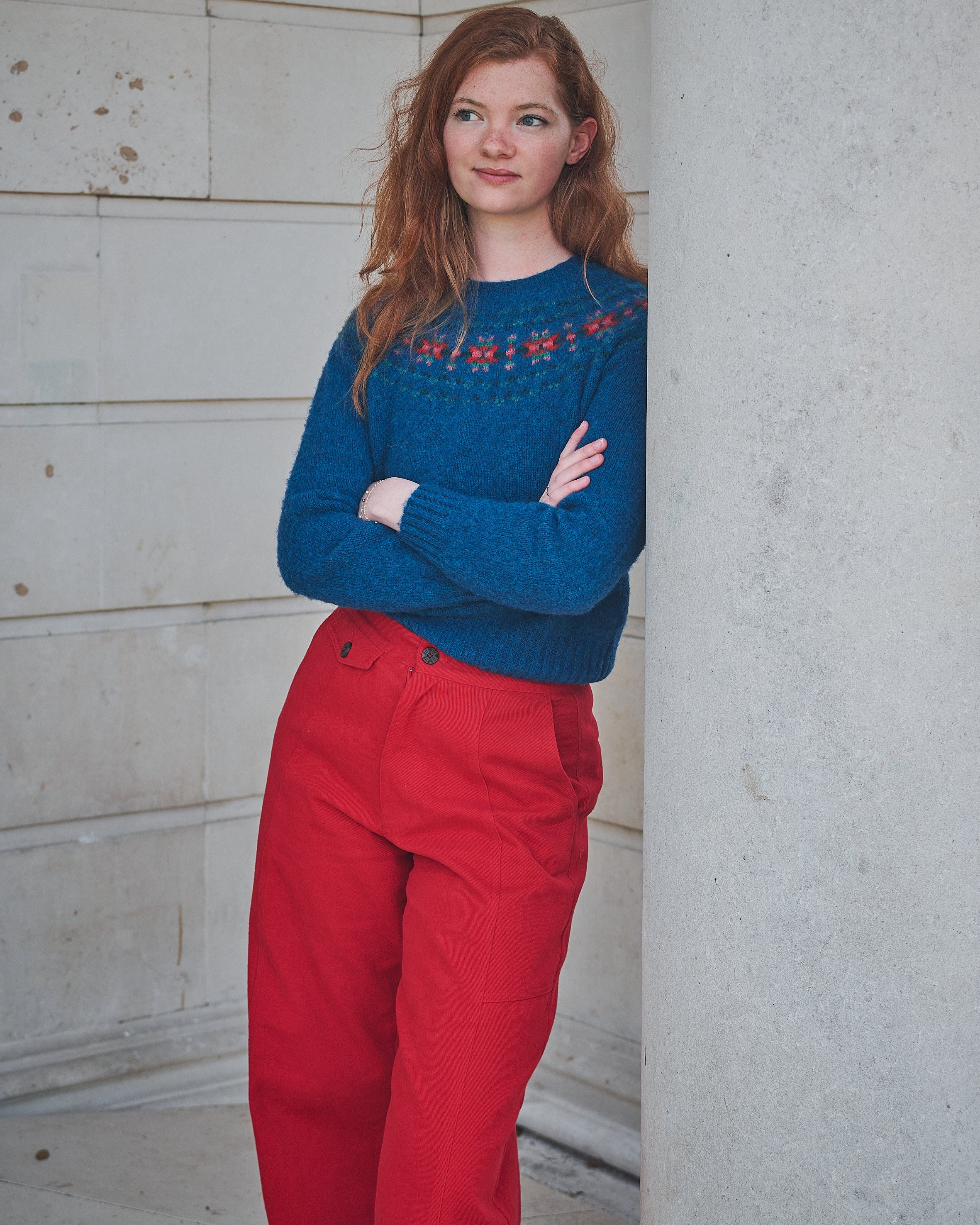 Woman wearing a blue sweater with red patterns and red pants standing against a light-colored wall.
