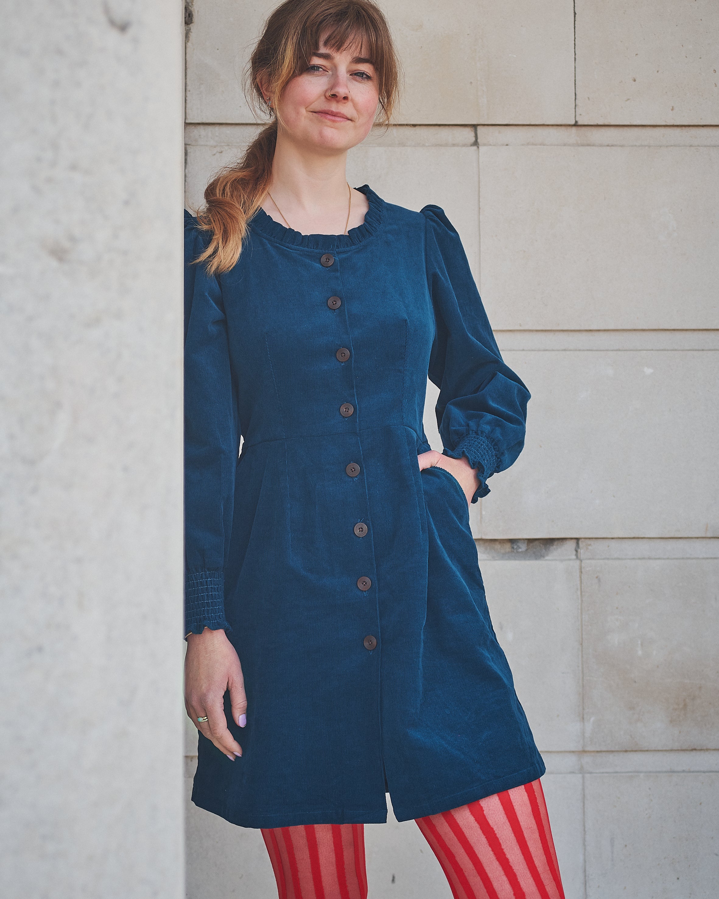 Woman wearing a blue dress with a button-up front, standing against a light stone wall.
