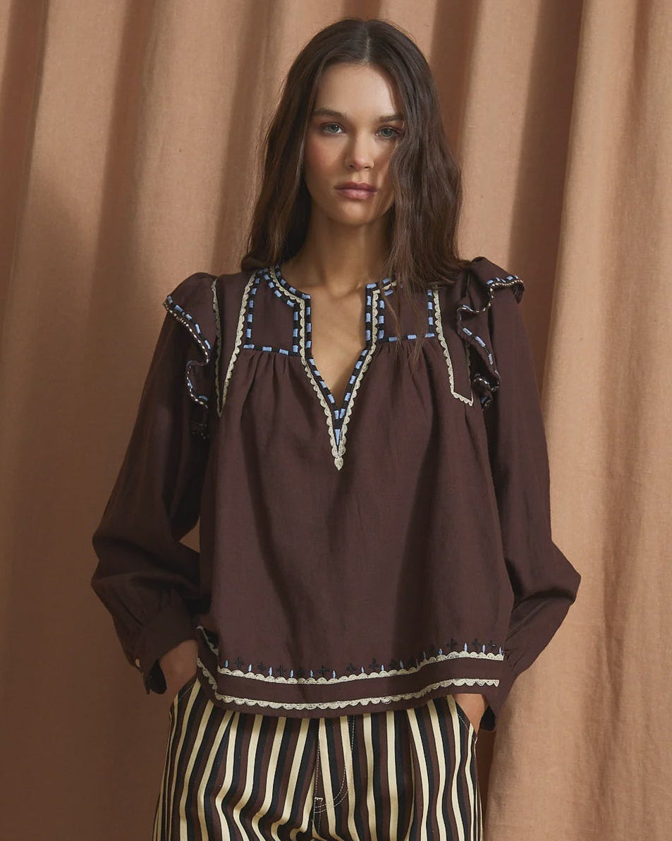 A woman modeling a dark brown long-sleeve blouse with embroidered detailing around the neckline and a striped black and cream wide-leg pant set, posed against a draped tan fabric backdrop.