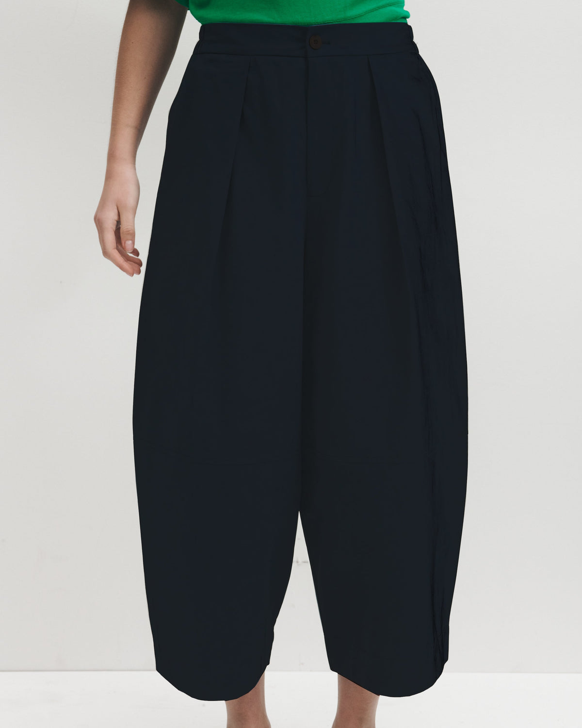 A model wearing high-waisted, voluminous dark navy pleated cropped trousers, styled with a bright green top and brown leather loafers against a plain white studio background.