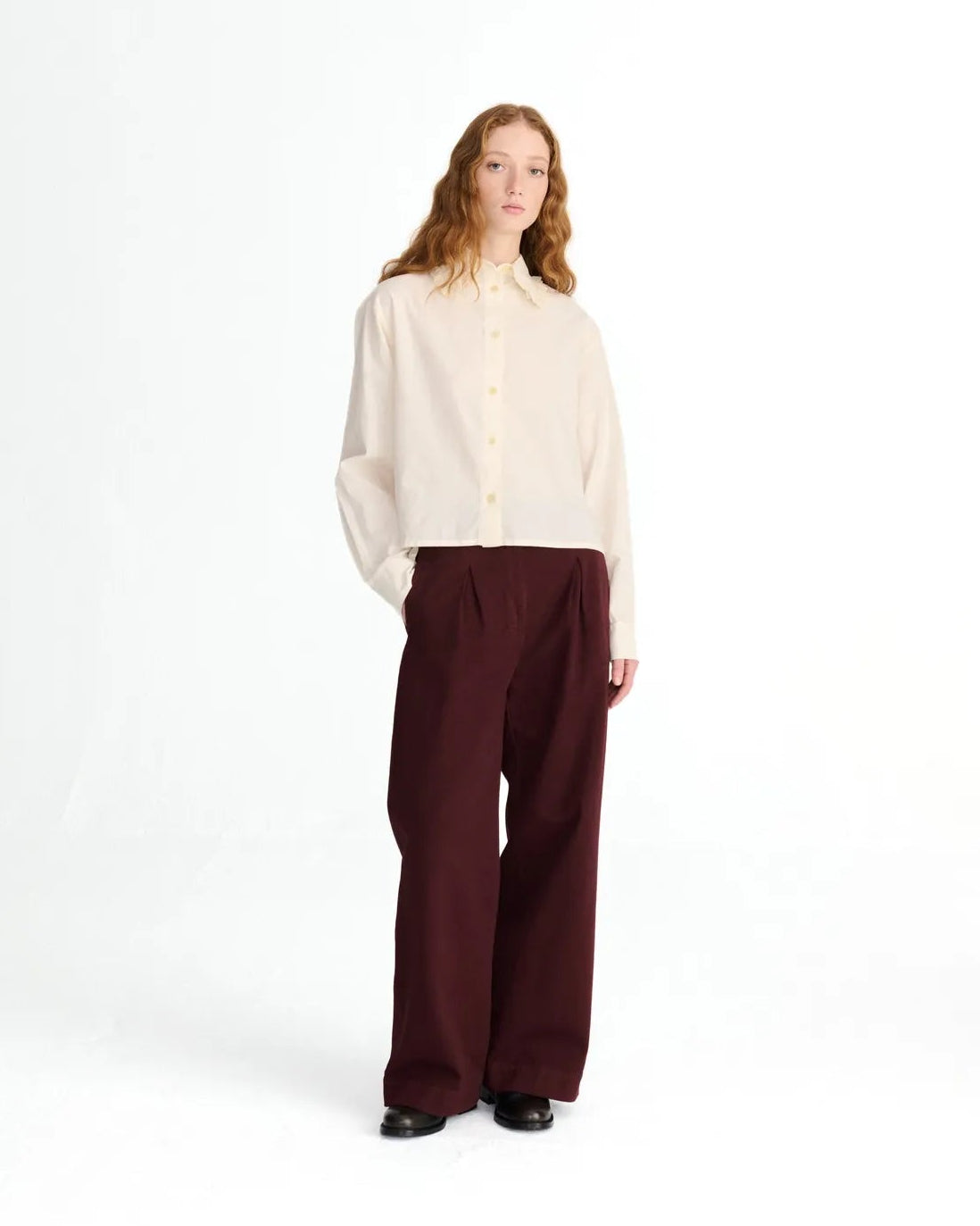 A model stands against a plain white studio background wearing a cream-colored long-sleeve button-up shirt and dark burgundy wide-leg trousers.