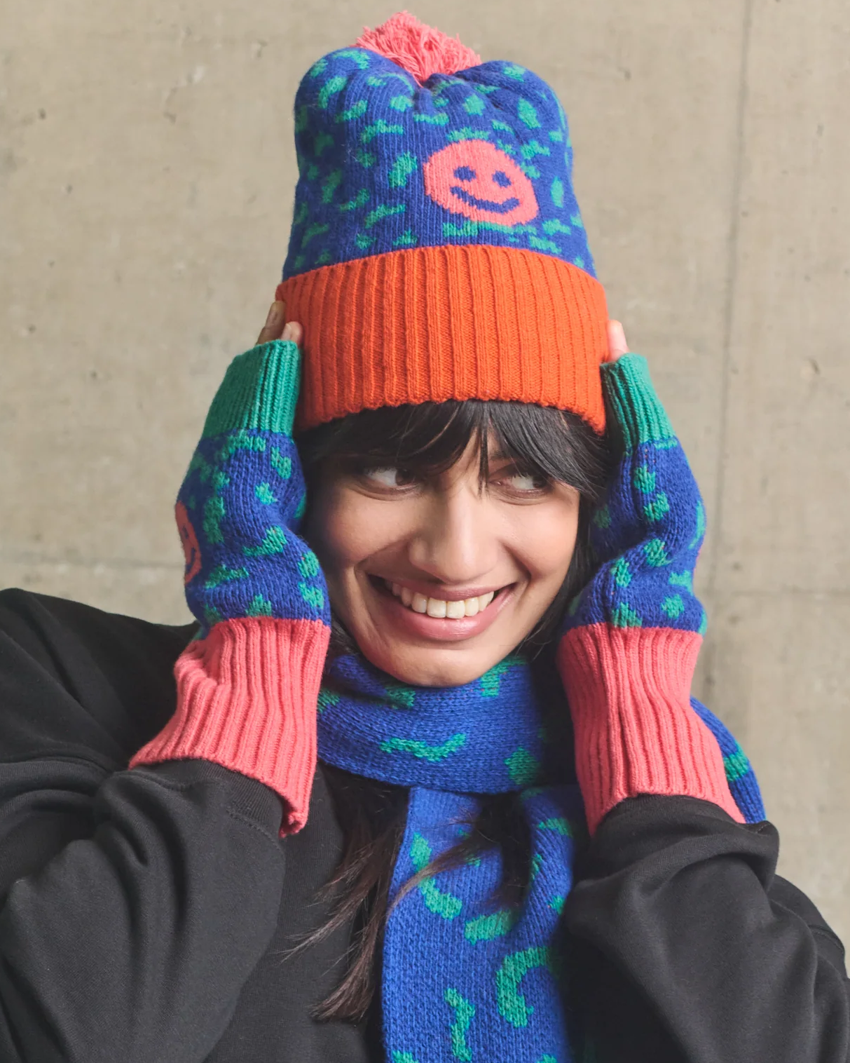 Person wearing a colorful knitted hat and scarf against a concrete wall.