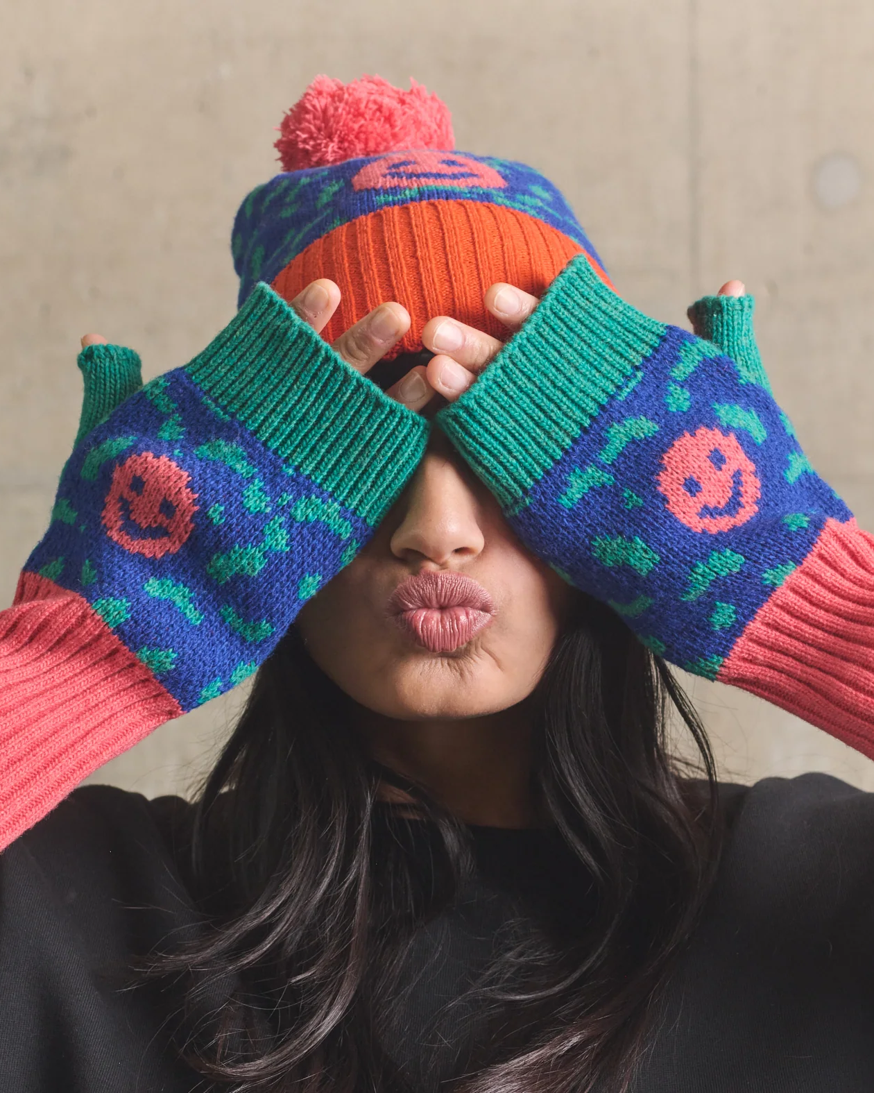 Person wearing colorful knitted hat and gloves against a concrete wall.