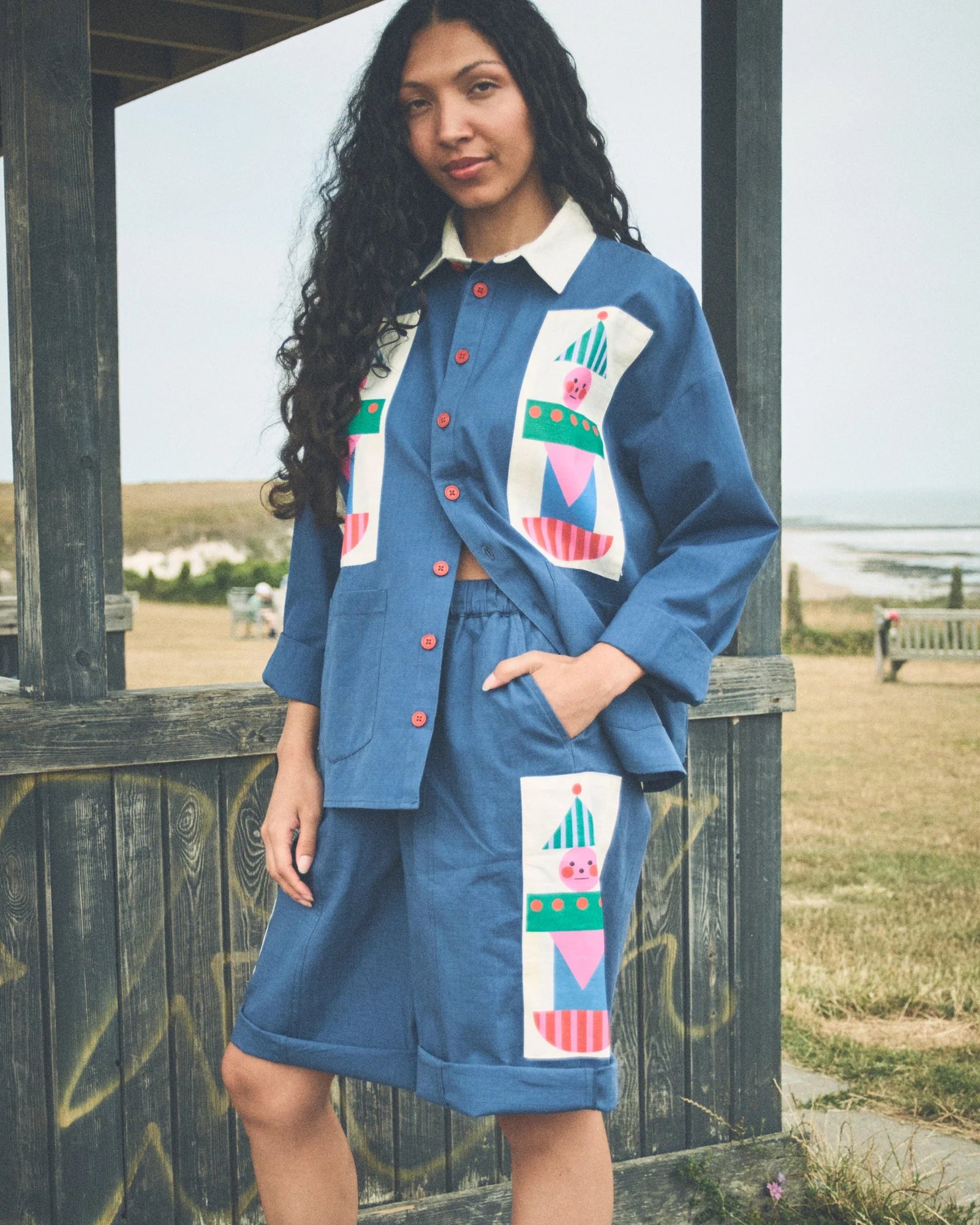 A woman stands outdoors in front of a wooden structure at a grassy coastal location, wearing a blue matching shirt and skirt set with large multicolor geometric patchwork panels on the front.