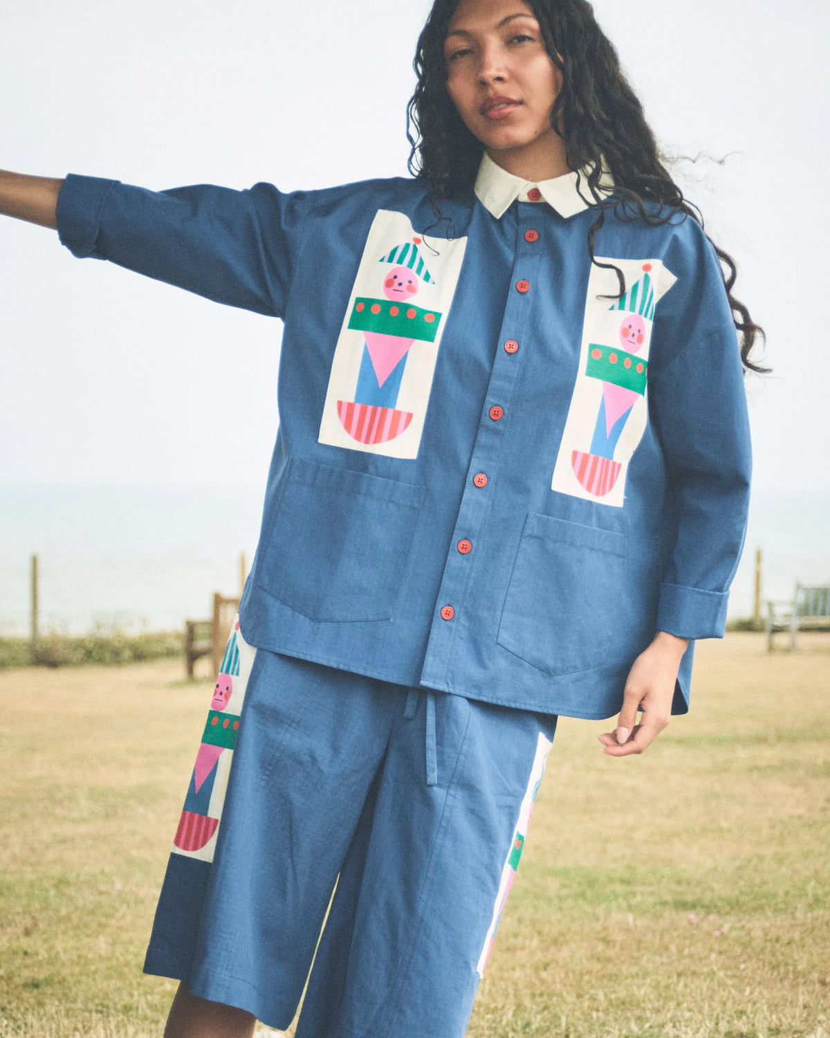 A woman wearing a blue denim-like matching set with colorful geometric patchwork details on the chest and sleeve, photographed outdoors near a grassy field and water.