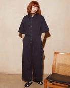 A model with red hair stands in a dark navy wide-leg jumpsuit with short sleeves and front buttons, standing next to a wooden cane chair in a minimalist room with a beige textured wall.