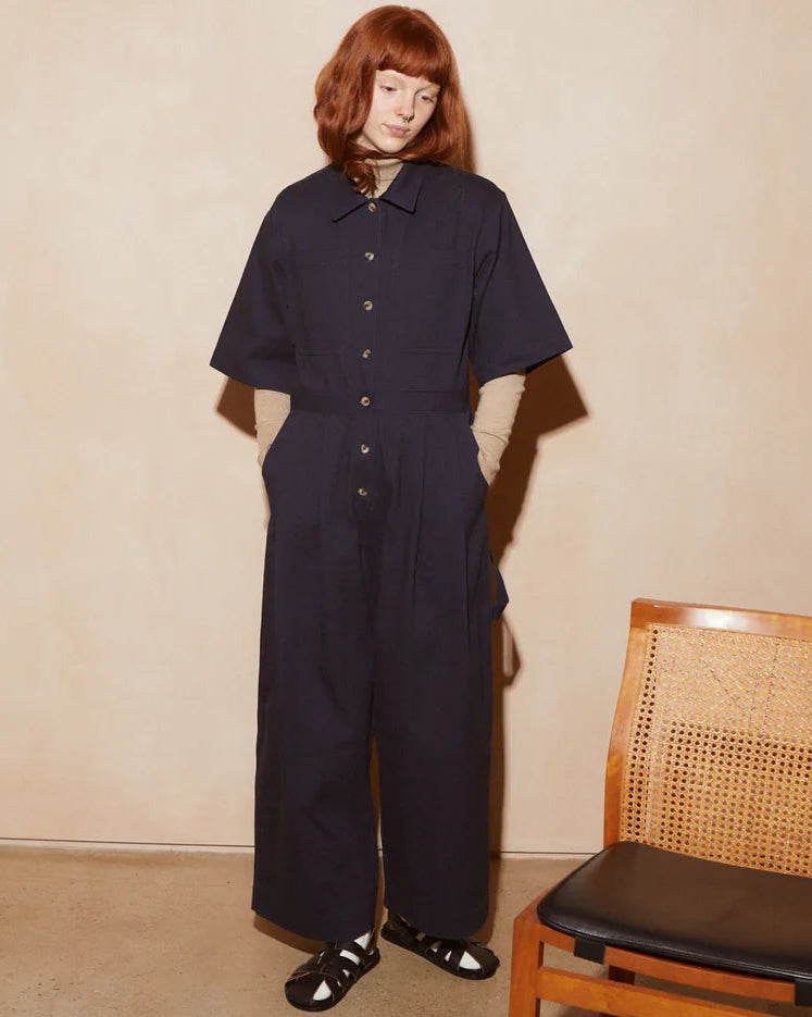 A model with red hair stands in a dark navy wide-leg jumpsuit with short sleeves and front buttons, standing next to a wooden cane chair in a minimalist room with a beige textured wall.