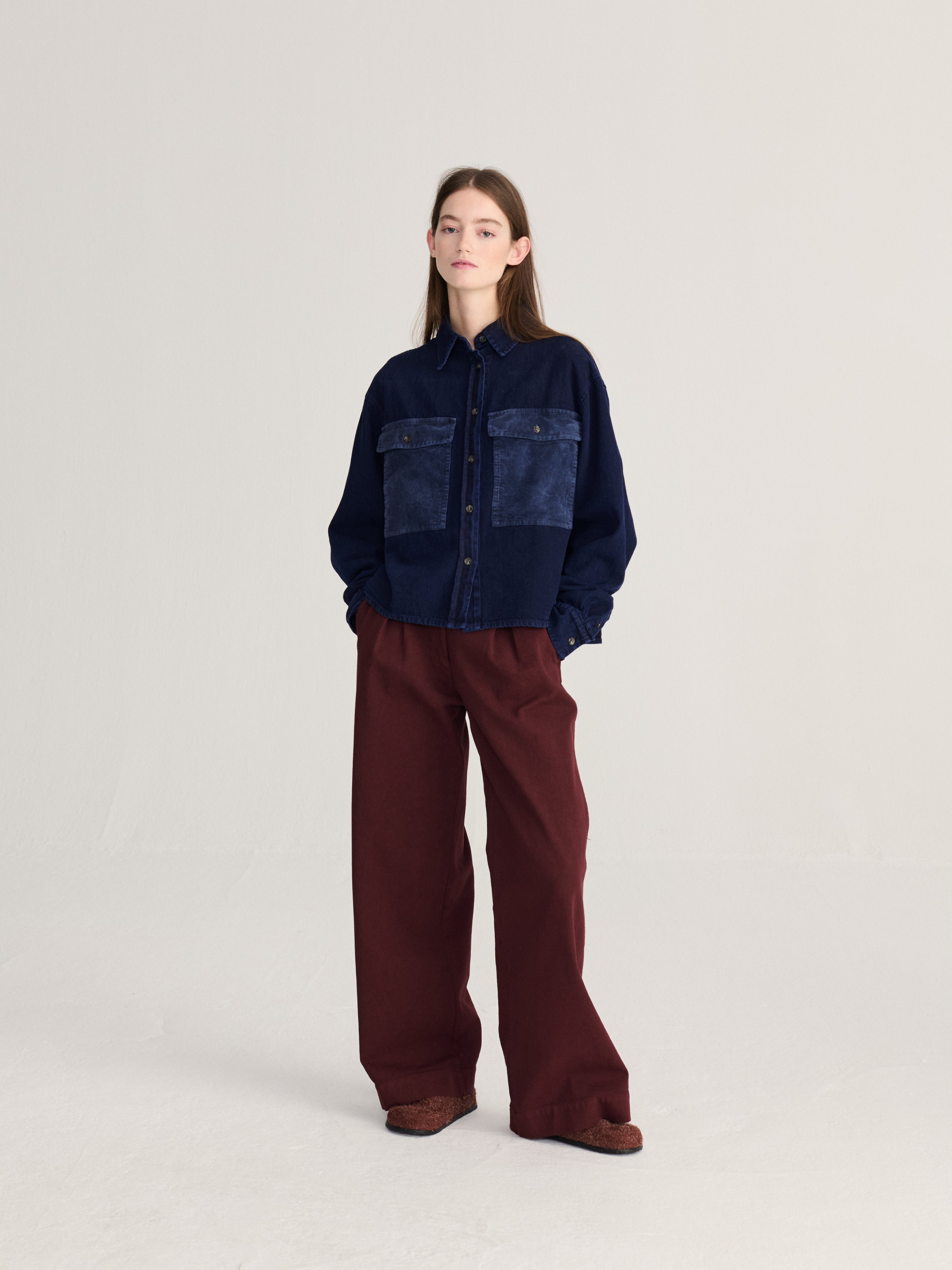 A model stands against a light gray studio background wearing a dark navy patchwork button-up shirt with wide-leg burgundy pants and brown shoes.