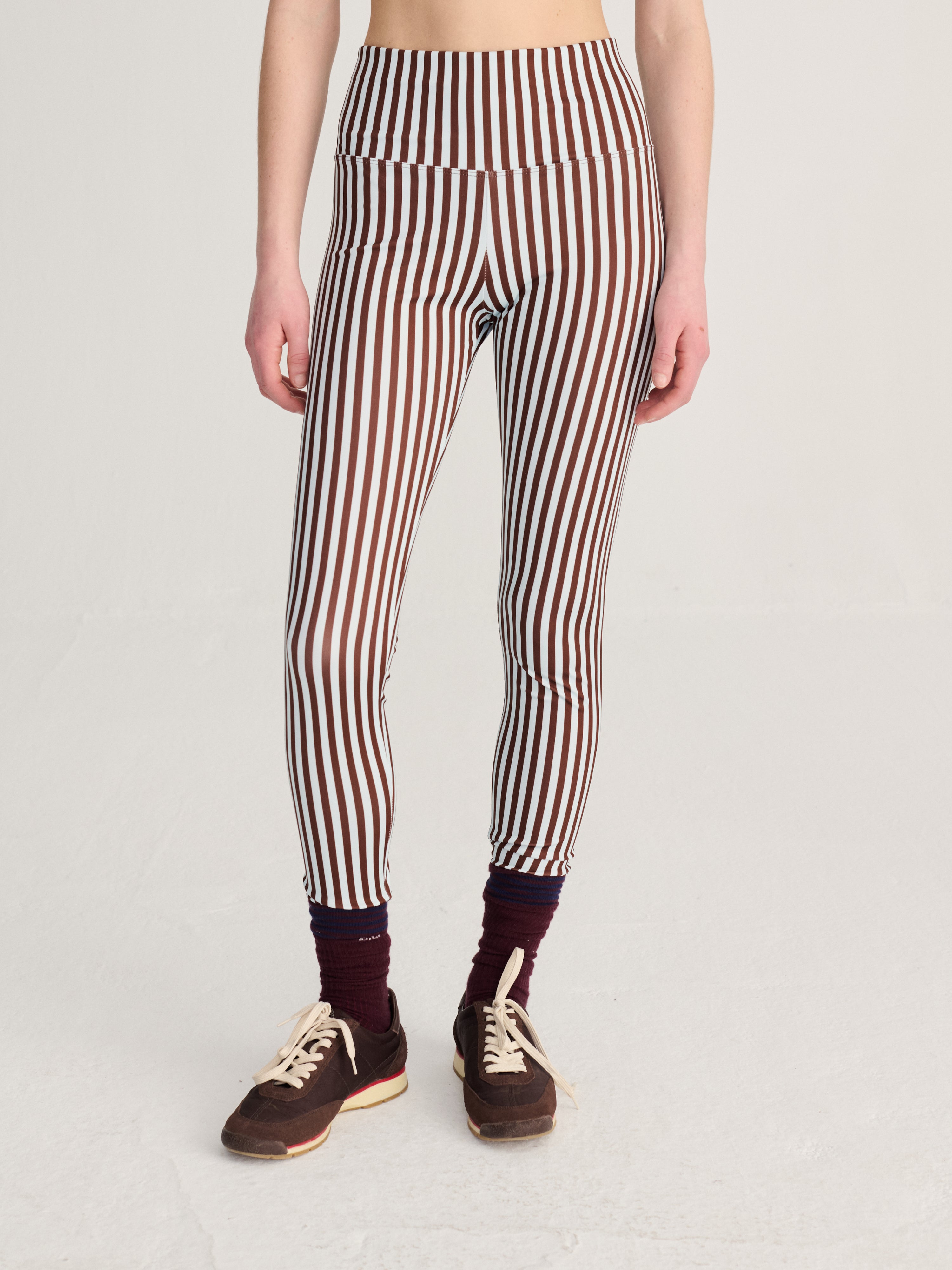 A model from the waist down wearing high-waisted leggings with vertical brown, white, and black stripes, styled with dark maroon socks and brown sneakers against a light gray studio background.
