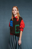 A model wearing a brown knit sweater vest with colorful abstract patches over a red long-sleeve shirt and a green, white, and black striped skirt, set against a textured dark blue-gray studio background.