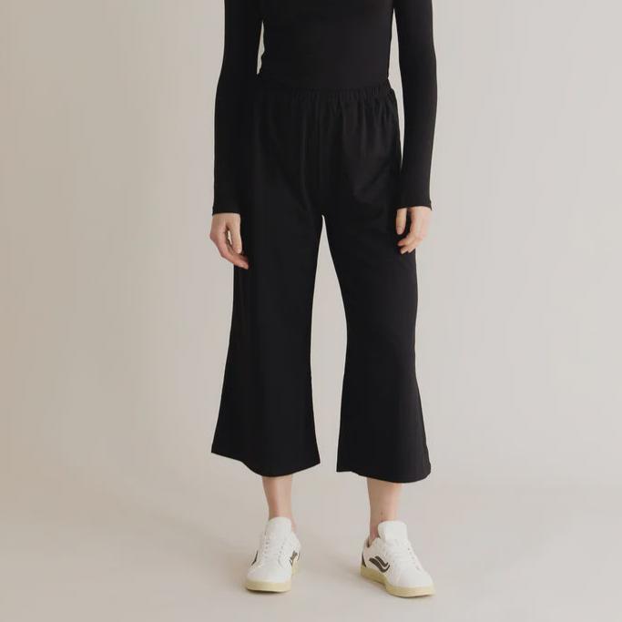 A model wearing black lounge pants with an elastic waistband and a relaxed fit. The pants are made from organic cotton and elastane, and the model is standing with one foot on a white shoe.